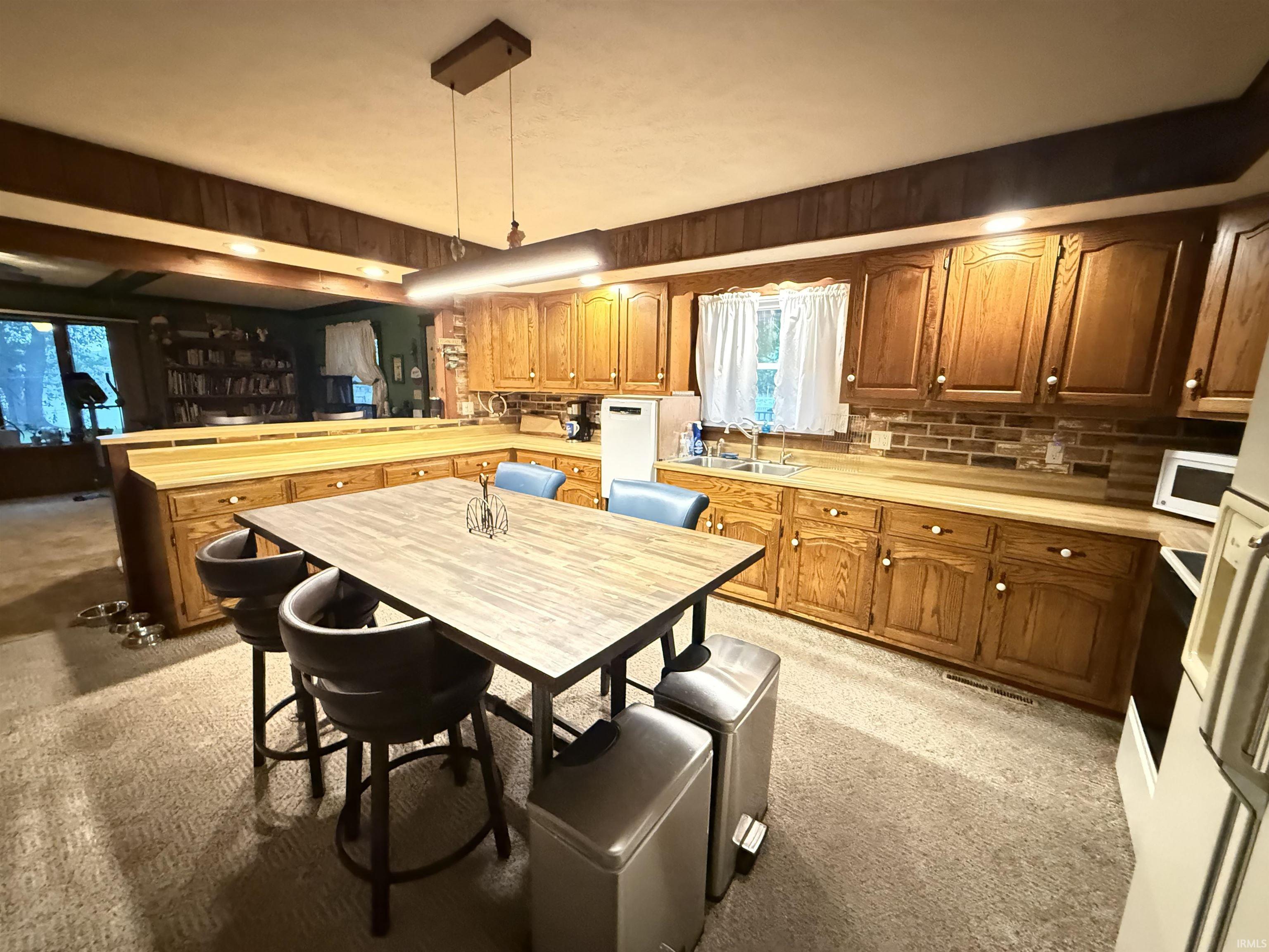 Wood cabinetry with white hardware, wood-finish countertops, stainless steel sink, brick-style tile backsplash, and neutral carpet flooring