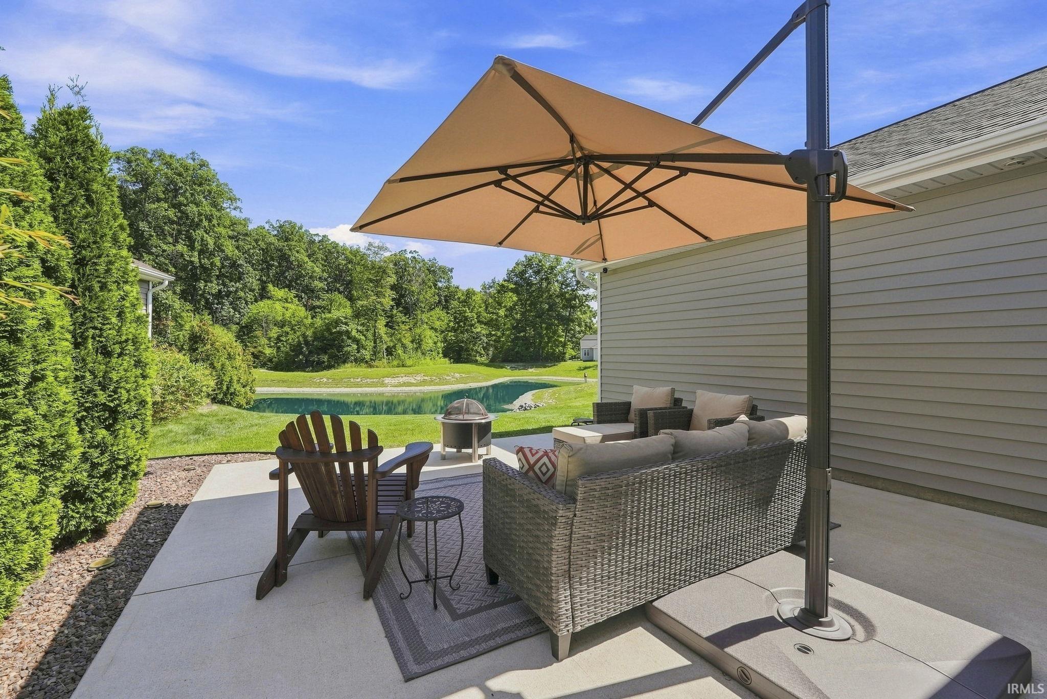 Expansive concrete patio with a detached umbrella, bordered by a manicured lawn, mature trees, and a visible pond