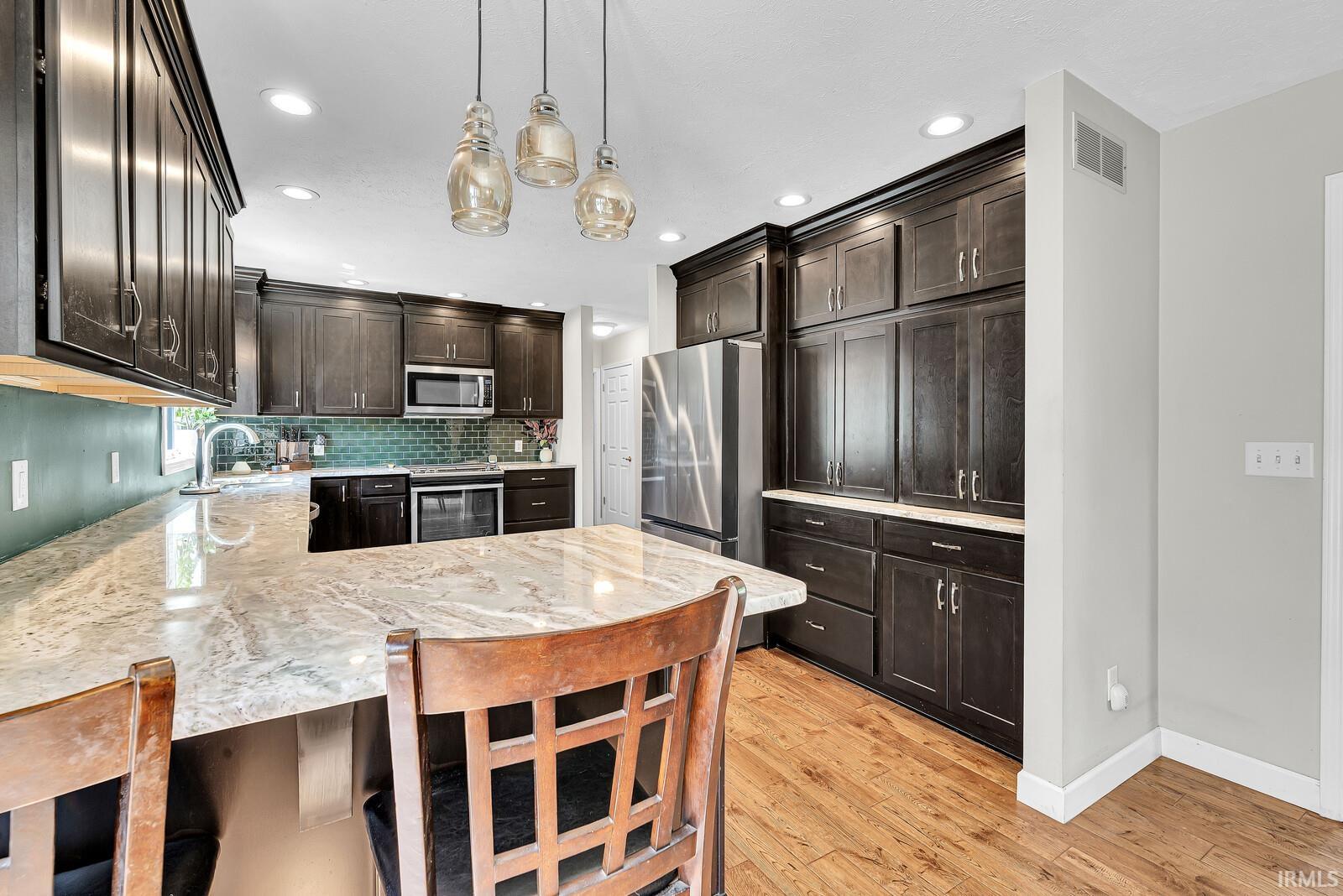 Kitchen featuring extensive dark wood-finish cabinetry, light-toned countertops, a multi-toned tile backsplash, stainless steel appliances, and wood-finish flooring