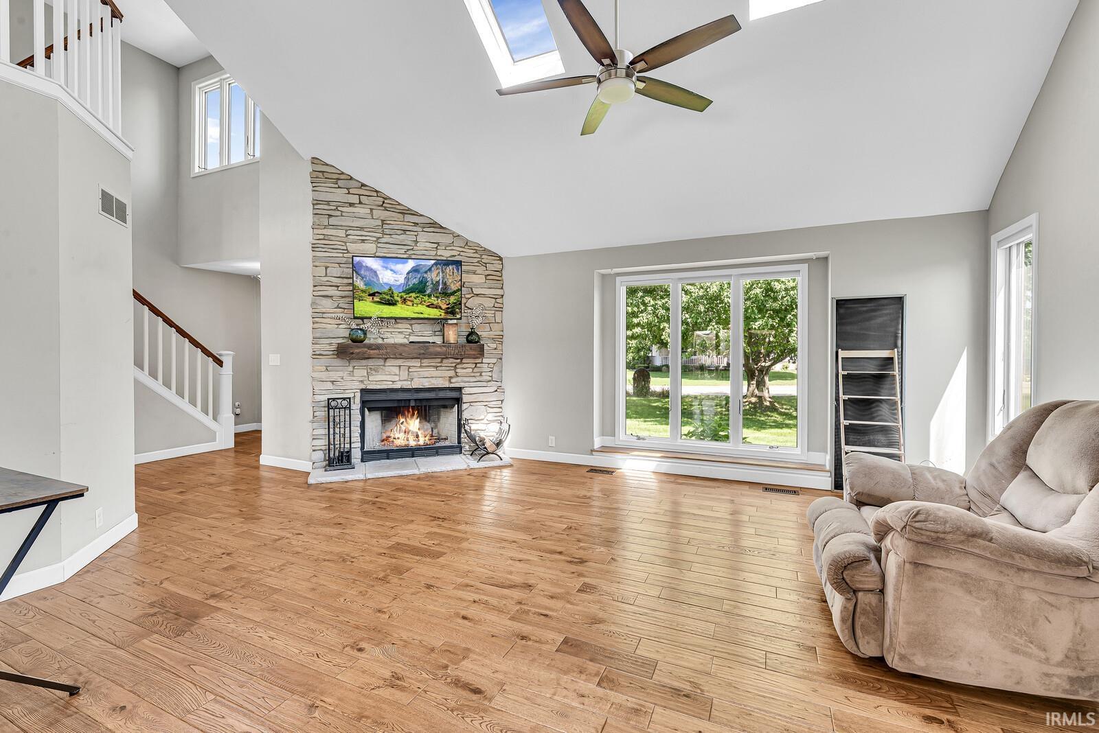 Vaulted ceiling featuring skylights and a ceiling fan, stone-clad fireplace with a wood mantel, wood-finish flooring, and large windows