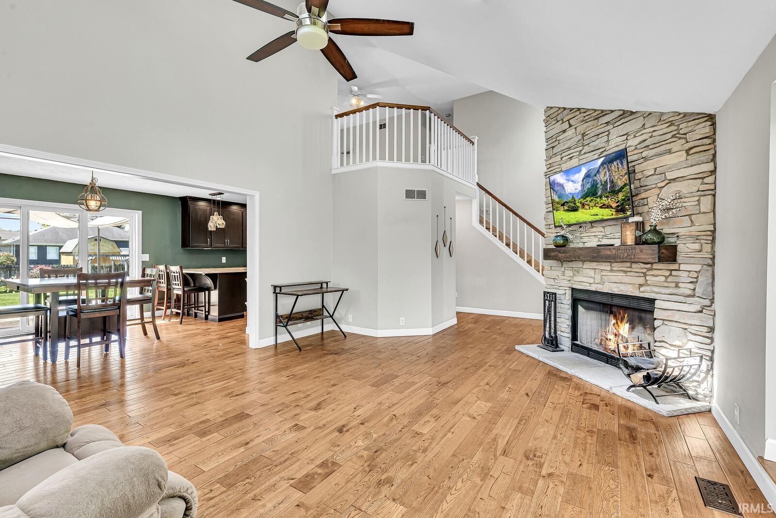 Vaulted ceiling with a ceiling fan, stone-clad fireplace with a wood mantle, wood-finish flooring, and a second-story interior balcony