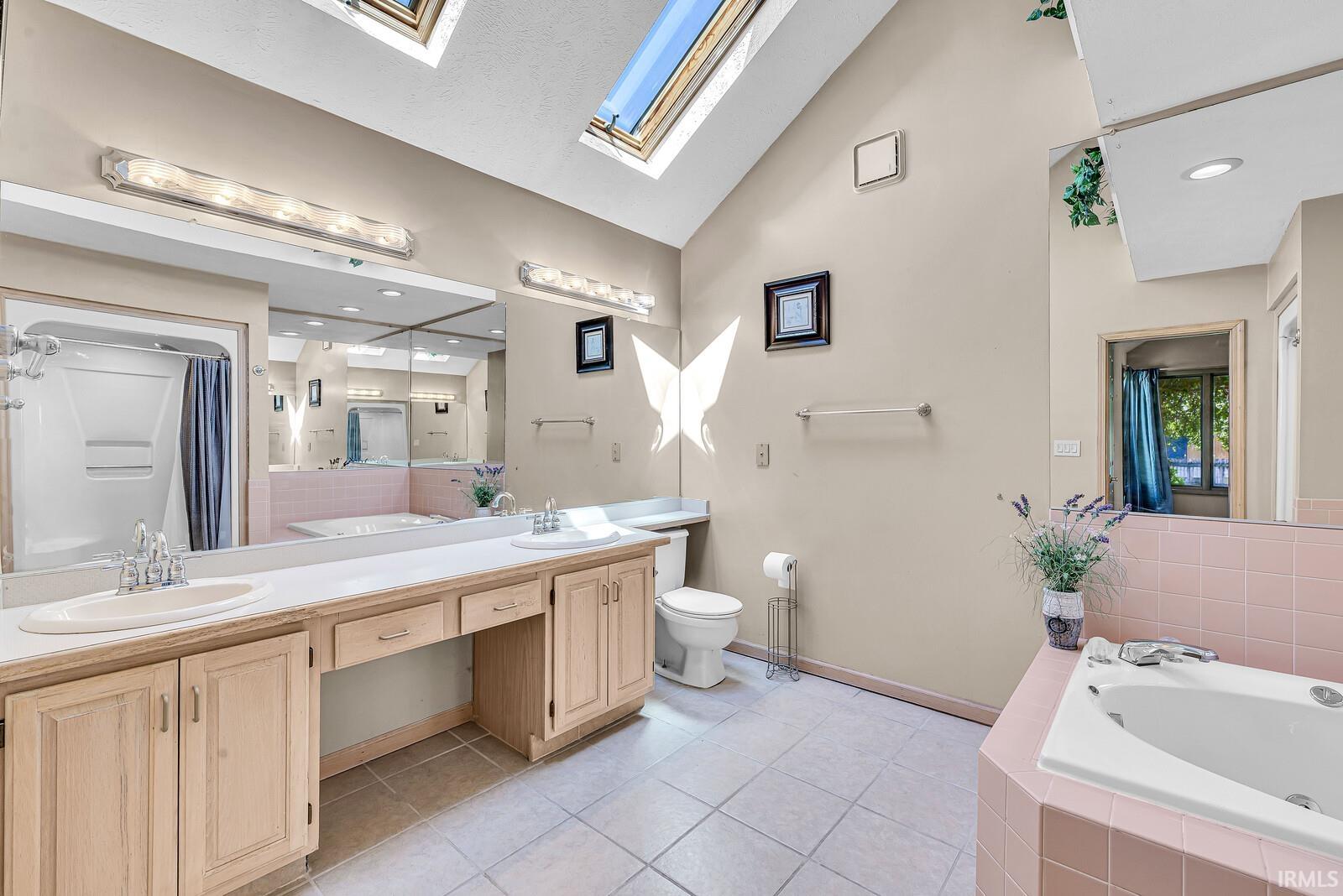 Vaulted ceiling with skylights, dual vanity with undermount sinks, large wall mirror, built-in soaking tub, and tile flooring