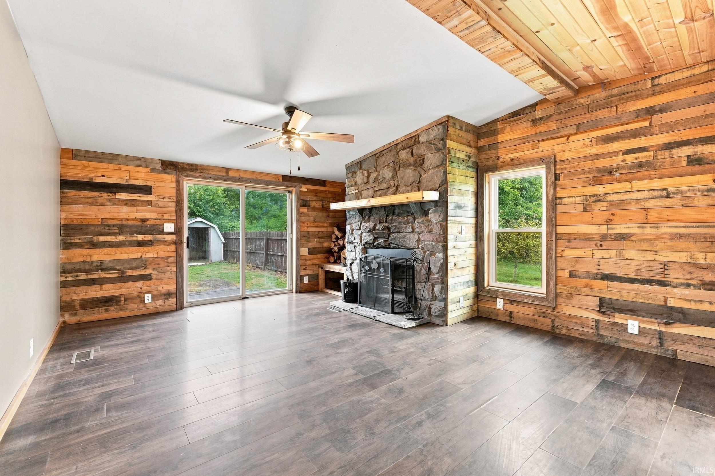 Wood-finish flooring, stone-clad fireplace with mantel, wood plank wall finishes, wood plank ceiling finishes, and a ceiling fan