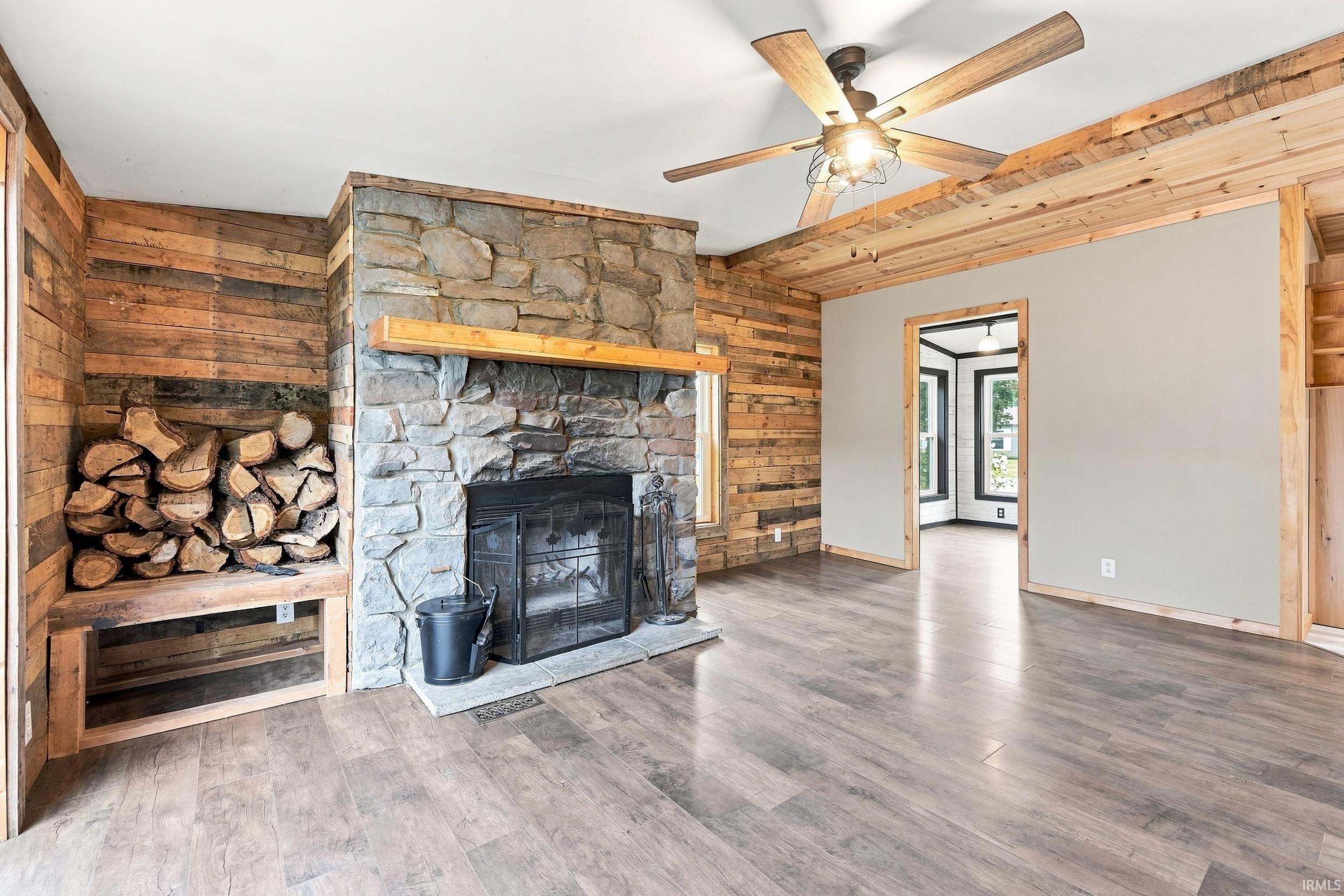 Stone fireplace with wood mantel, wood-finish flooring, wood plank accent walls, wood-beamed ceiling, and a ceiling fan with light fixture