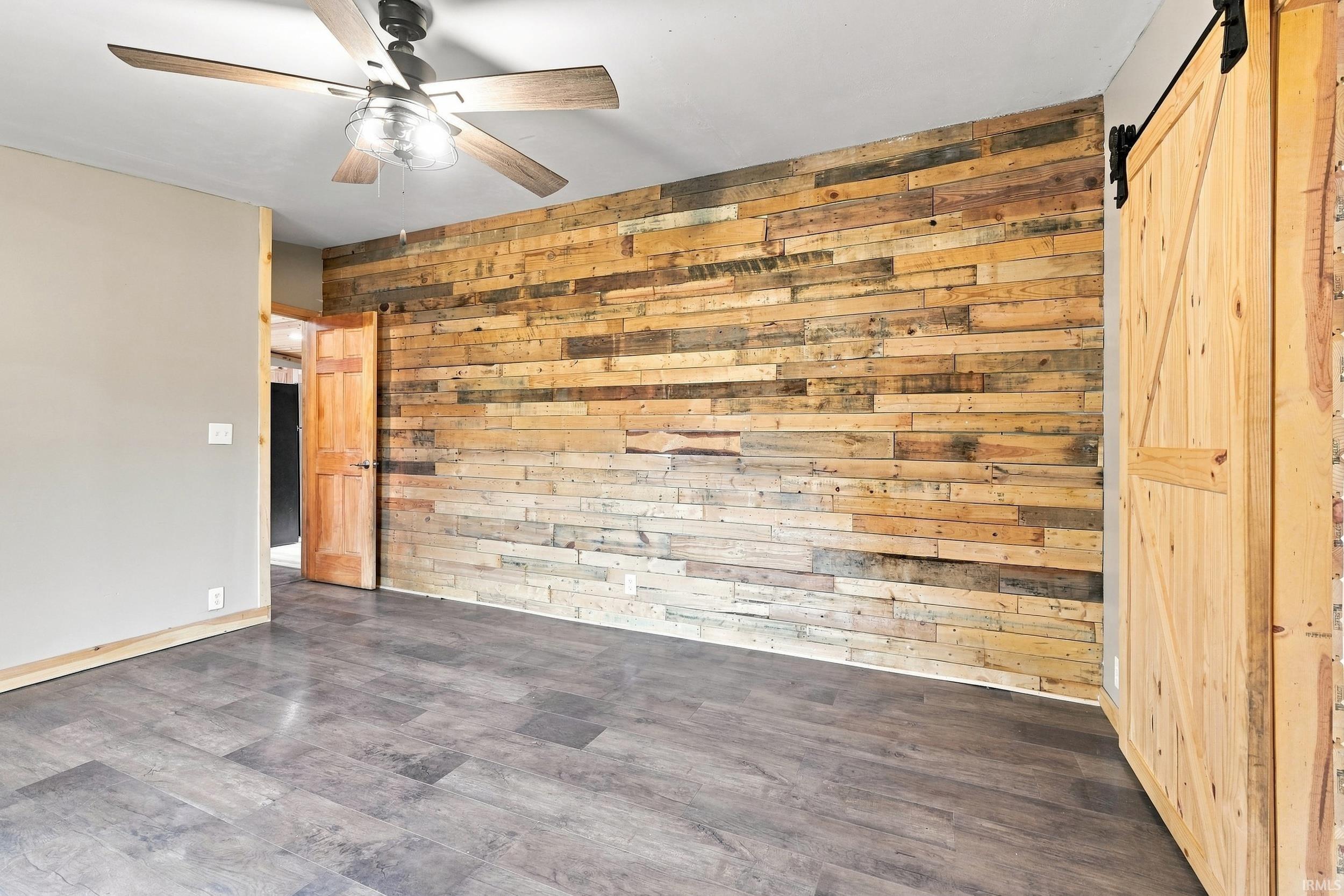 Wood-finish flooring, accent wall with wood paneling, natural wood barn door, and a ceiling fan with integrated lighting