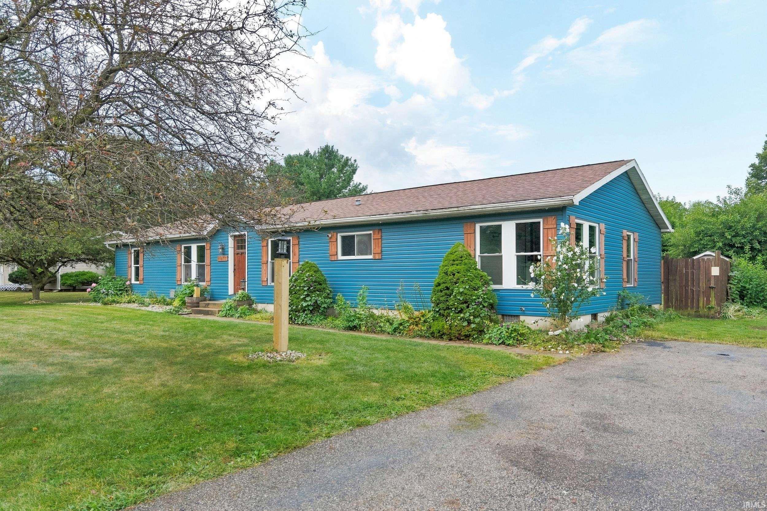 Blue exterior siding with white trim, brown shingle roof, wood-tone shutters, mature landscaping, and a paved driveway