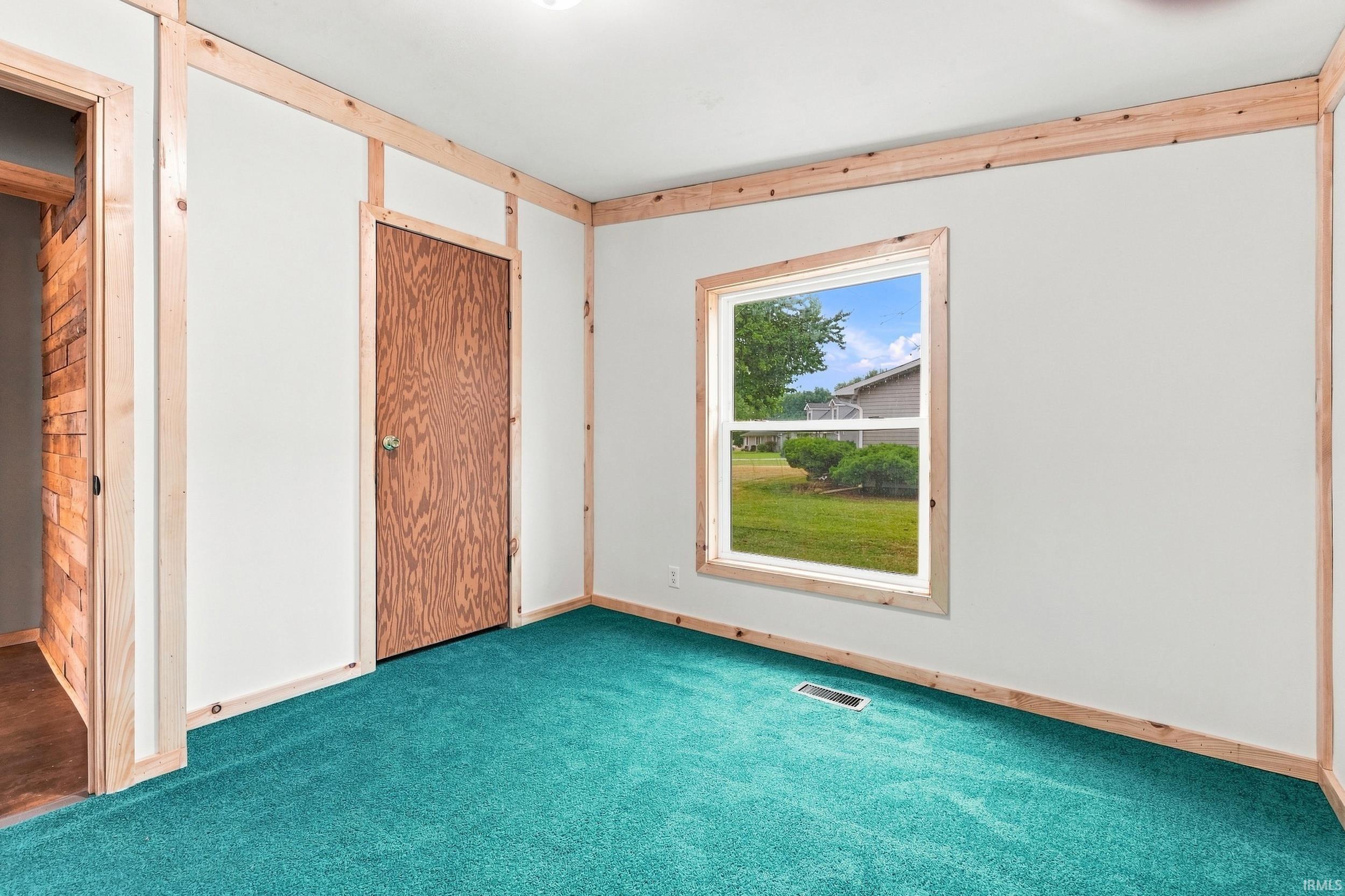 White walls with natural wood trim, a single window, a wood-finish door, and teal carpeting