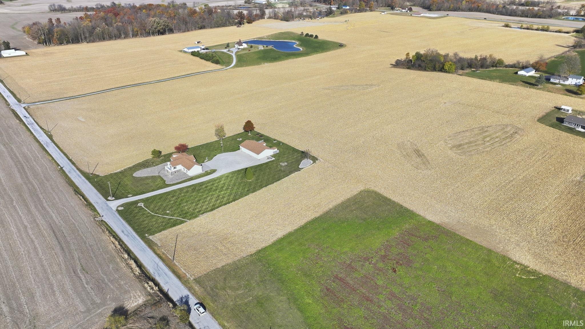 Aerial perspective of expansive acreage featuring cultivated crop land, a private pond, a paved driveway, and manicured grounds surrounding the primary structure