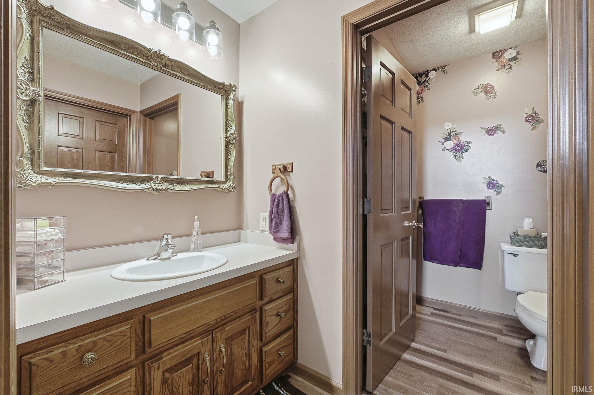 Wood-finish vanity cabinet with a solid-surface countertop and integrated basin, decorative wall-mounted mirror, and wood-finish flooring