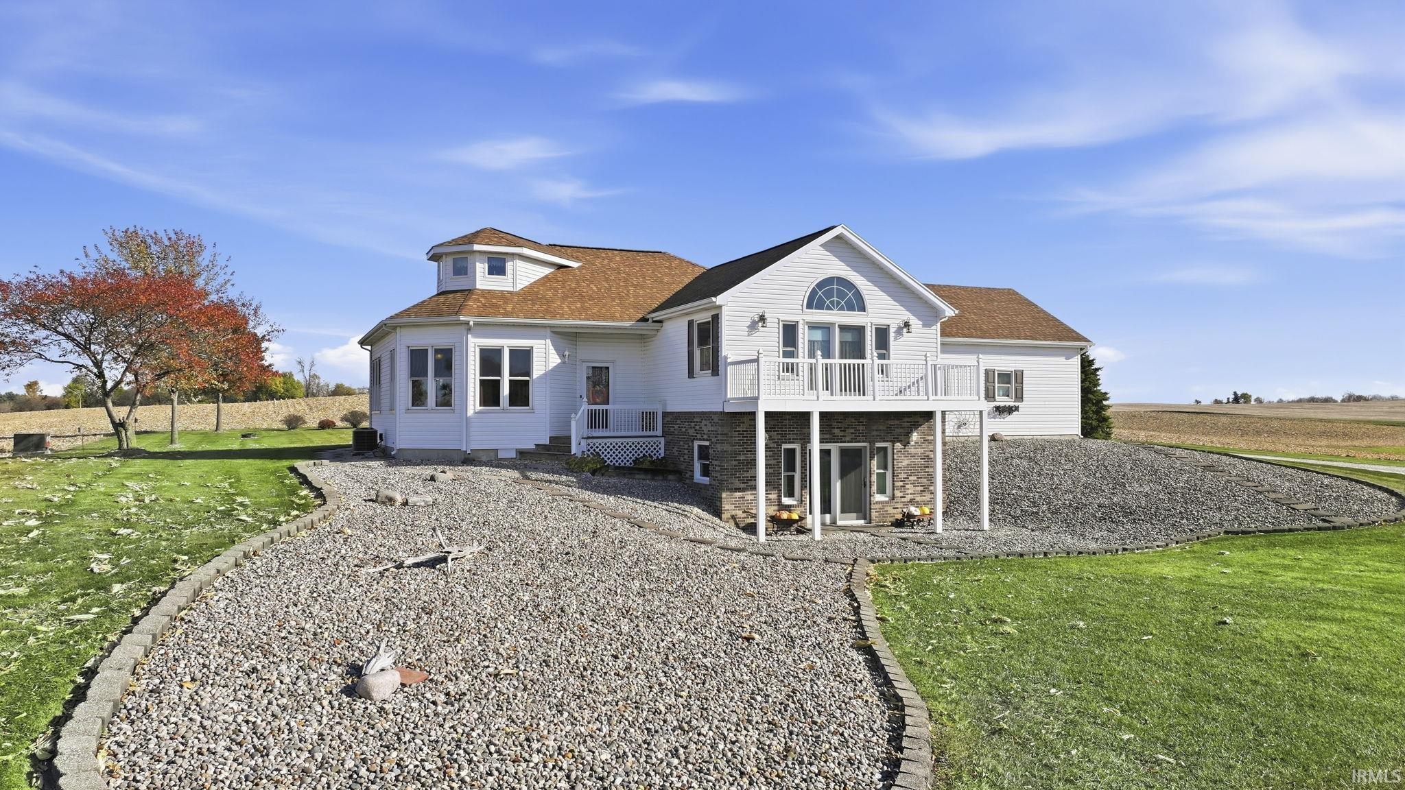 Two-story residence featuring white siding, a multi-faceted roofline with turret detail, stone masonry foundation, an elevated deck with white railing, and arched window accent