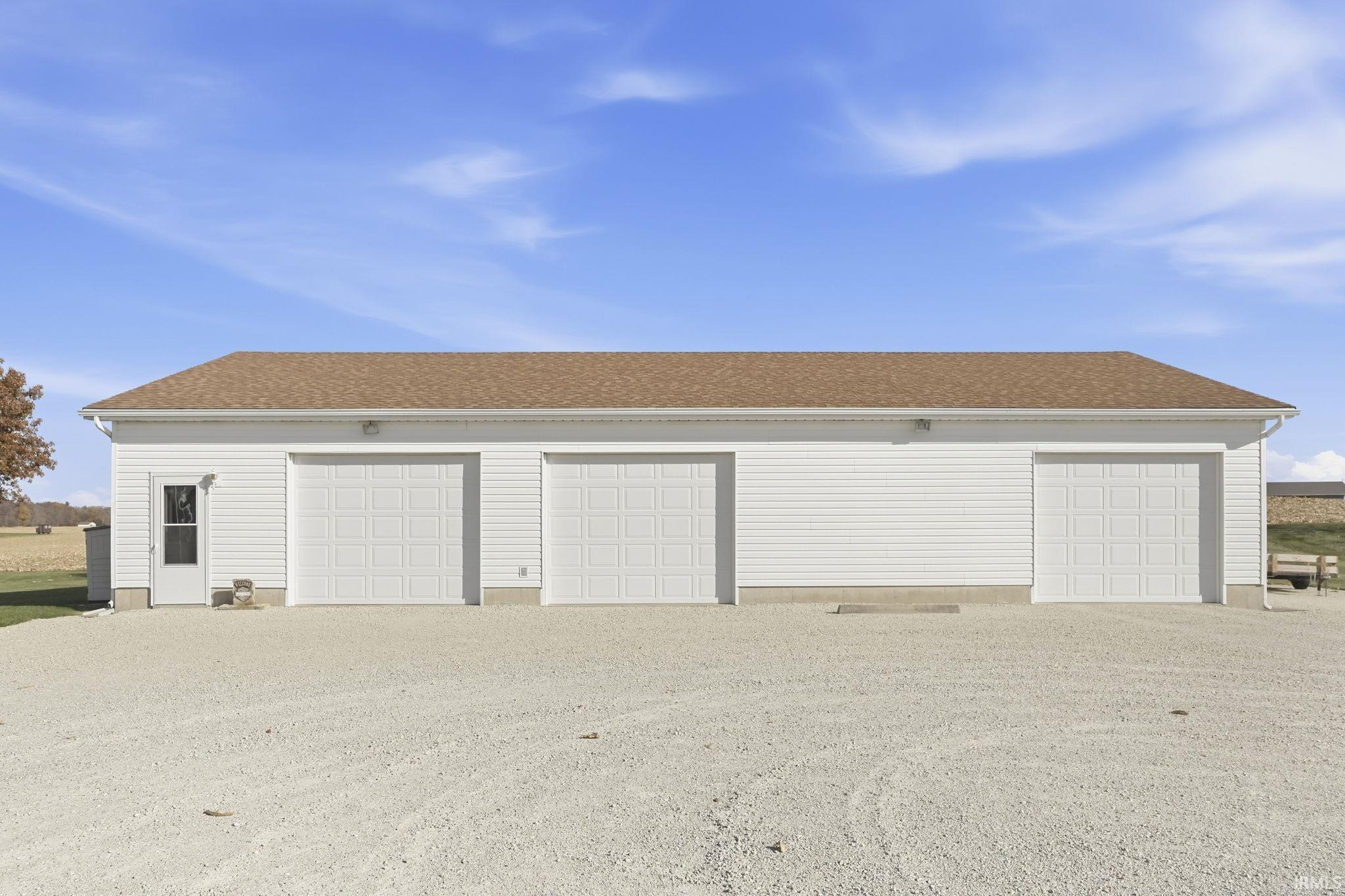 Detached three-car garage featuring white vinyl siding, a brown shingled roof, three sectional overhead doors, and an exterior pedestrian door