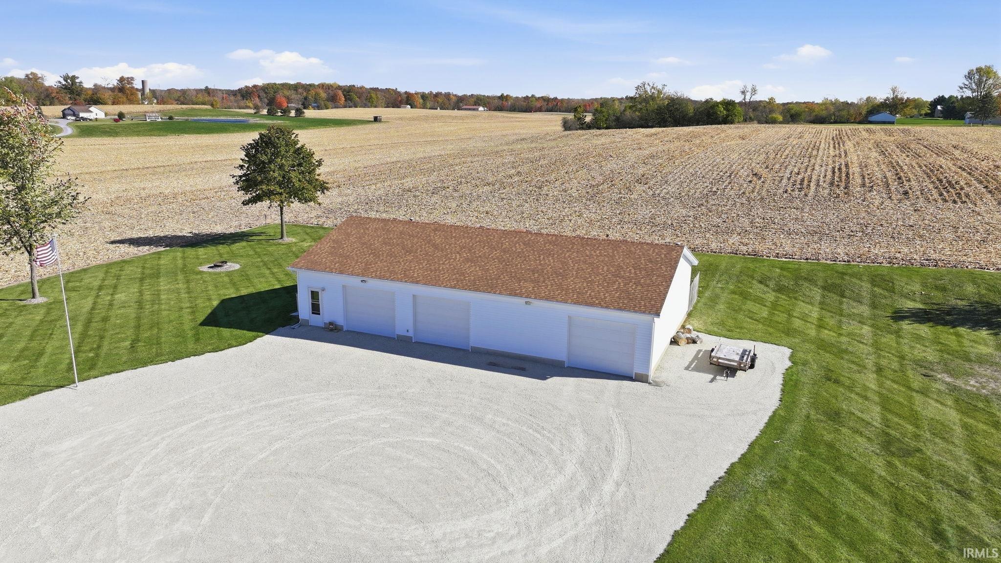 Detached three-car garage featuring a shingle roof, white siding, and a large gravel parking area situated on expansive acreage