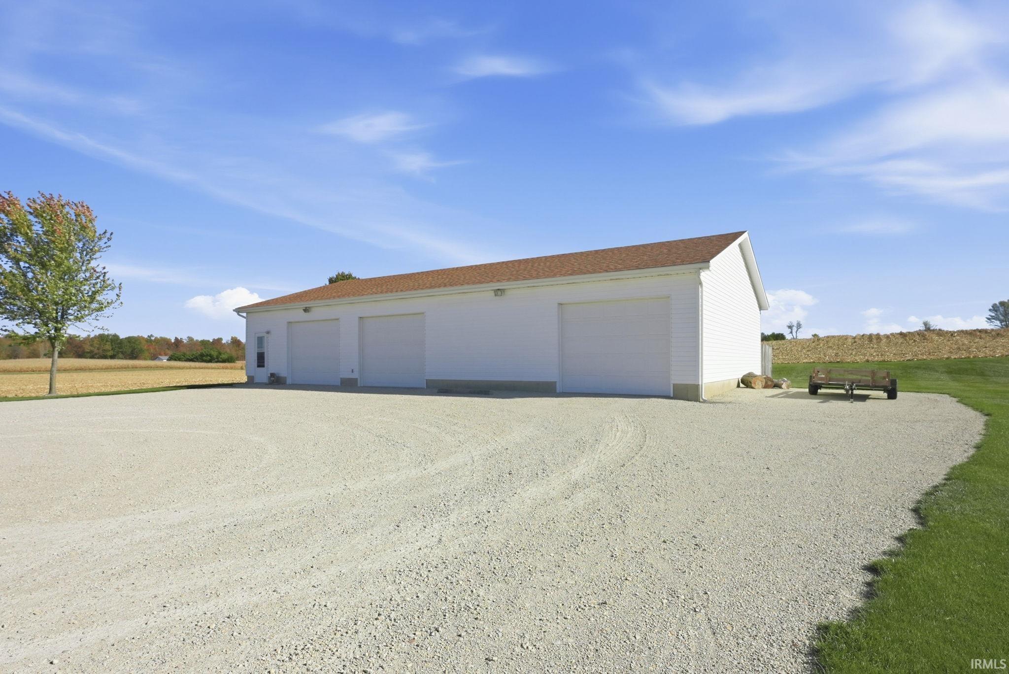 Detached three-bay garage featuring white siding, a brown shingled roof, and an expansive gravel driveway
