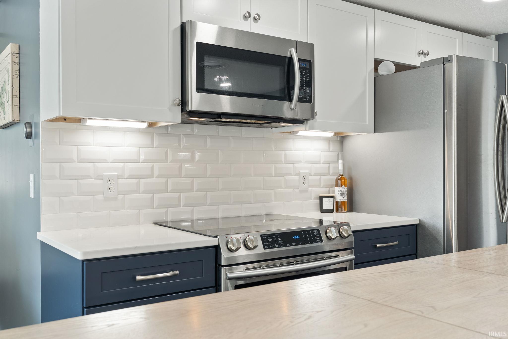 White subway tile backsplash, white upper cabinetry, stainless steel appliances, blue lower cabinetry, and light-toned countertops