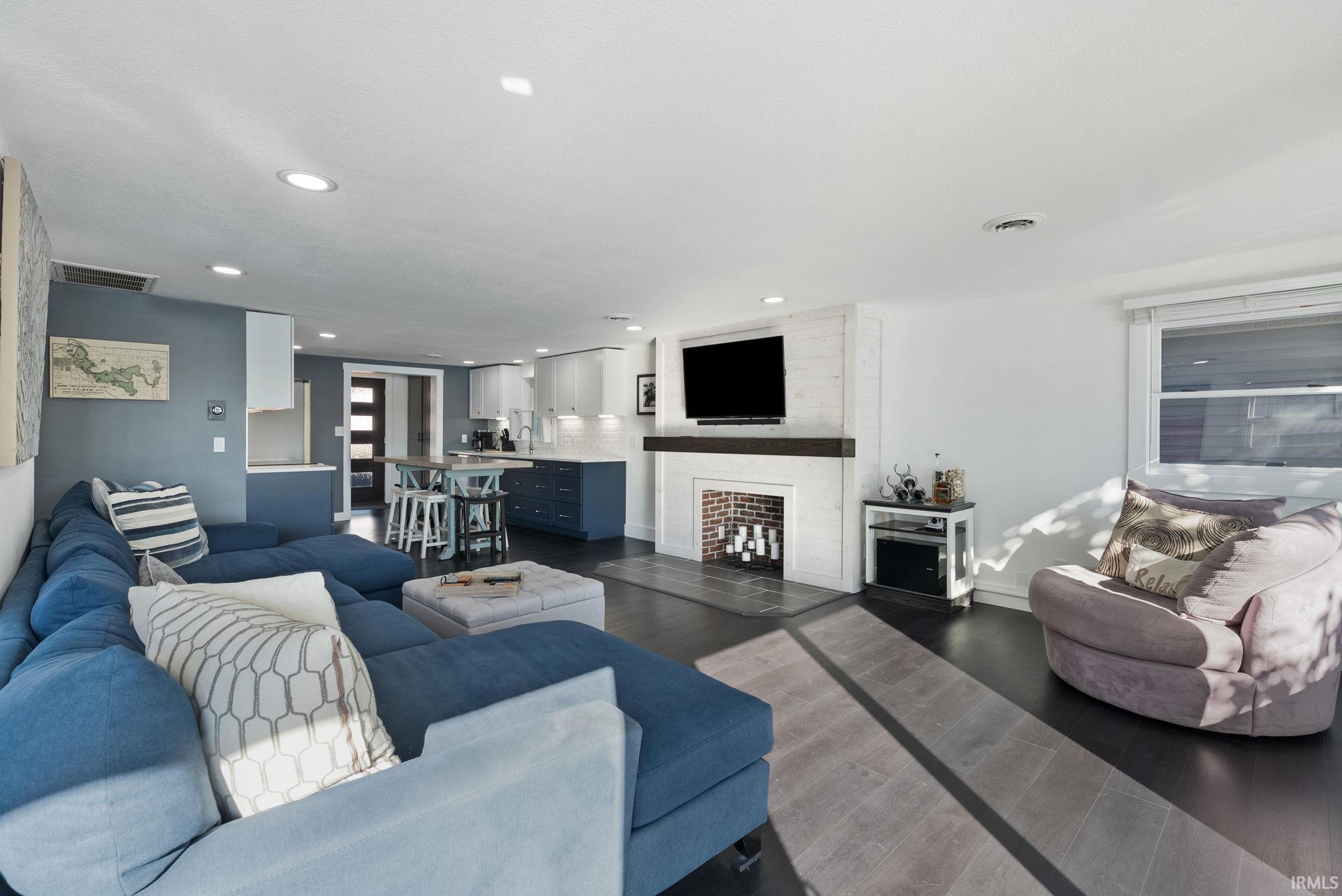 Open-concept living space featuring wood-finish flooring, recessed lighting, and a white brick-surround fireplace with a wood mantel