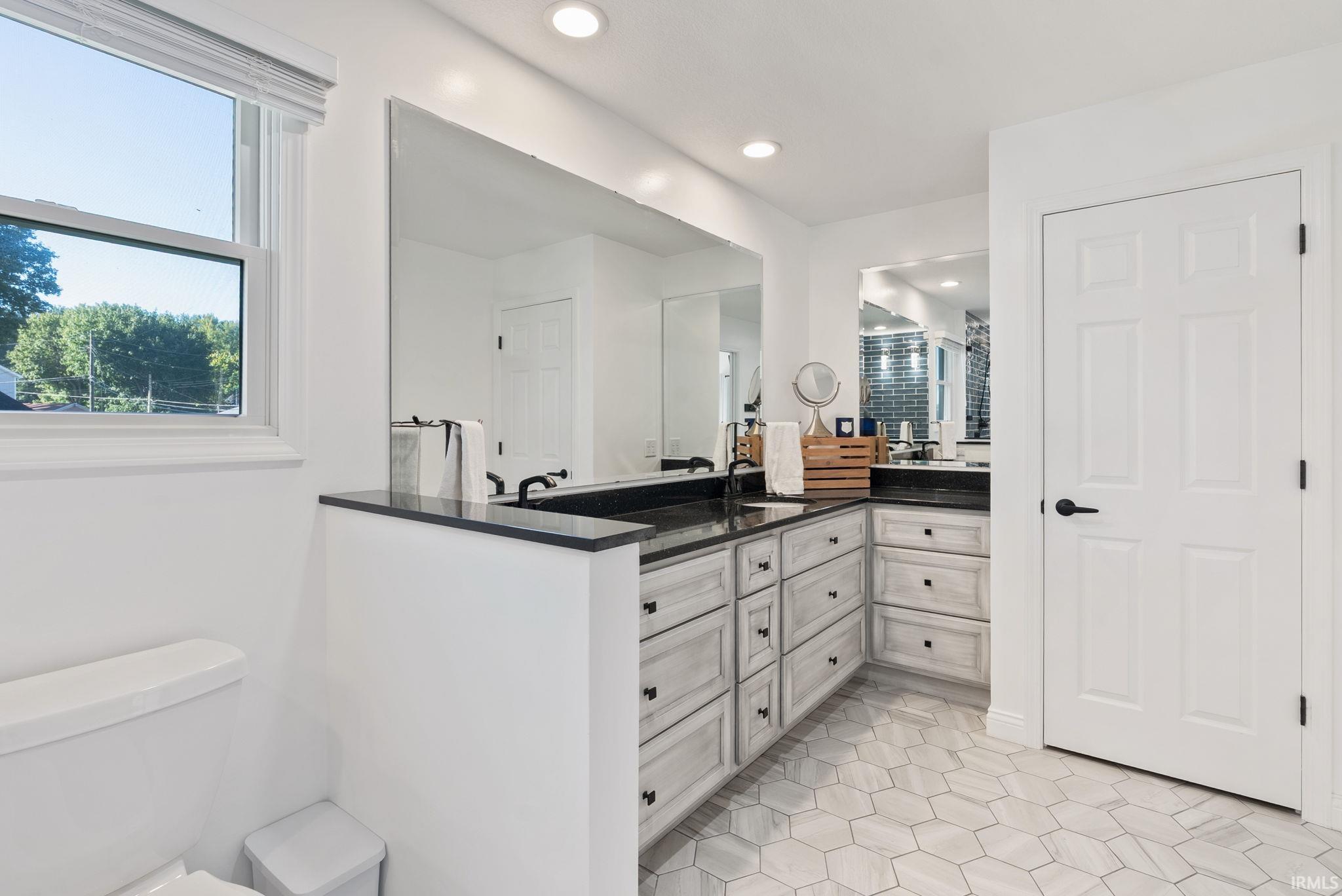 White hexagonal floor tiling, dual vanity with dark countertops and integrated sinks, extensive cabinetry storage, and a large wall-mounted mirror