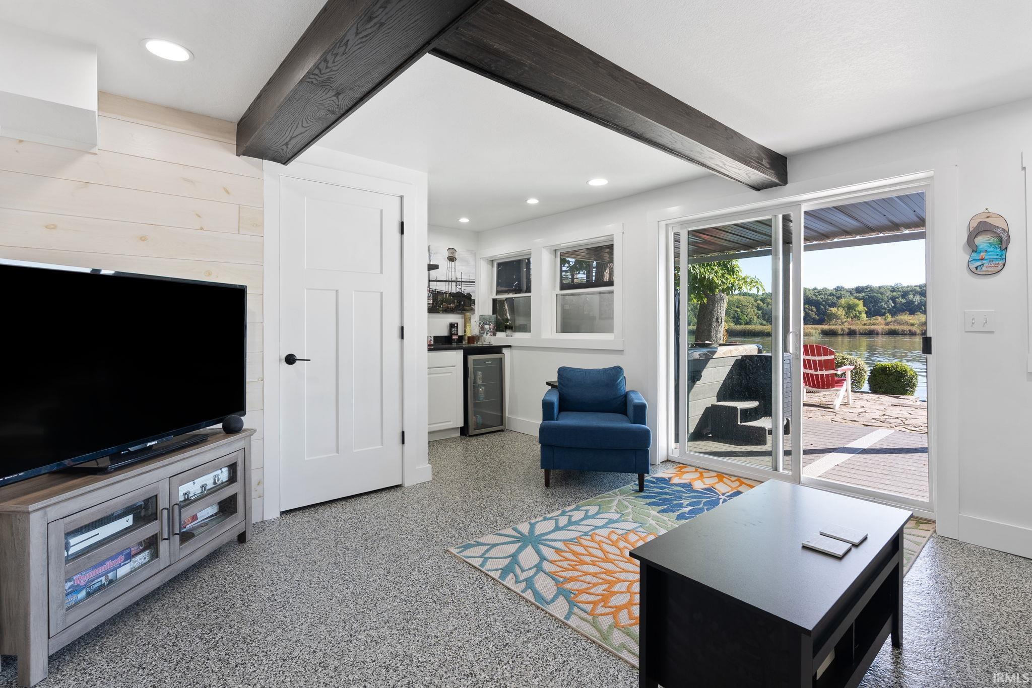 White shiplap accent wall, exposed dark wood ceiling beams, recessed lighting, and a kitchen area with built-in cabinetry