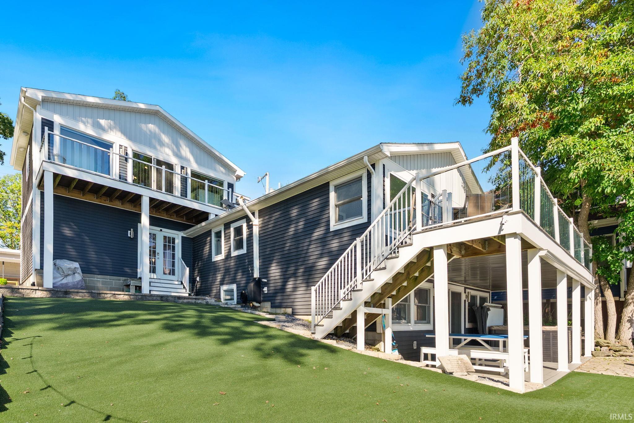 Expansive artificial turf lawn, multi-level decking with glass panel railings, and a lower-level patio with overhead deck coverage