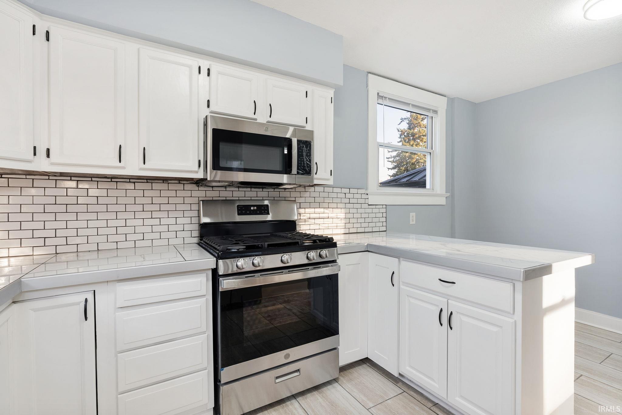 Kitchen featuring white cabinetry, stainless steel gas range and microwave, subway tile backsplash, and light-toned tile flooring