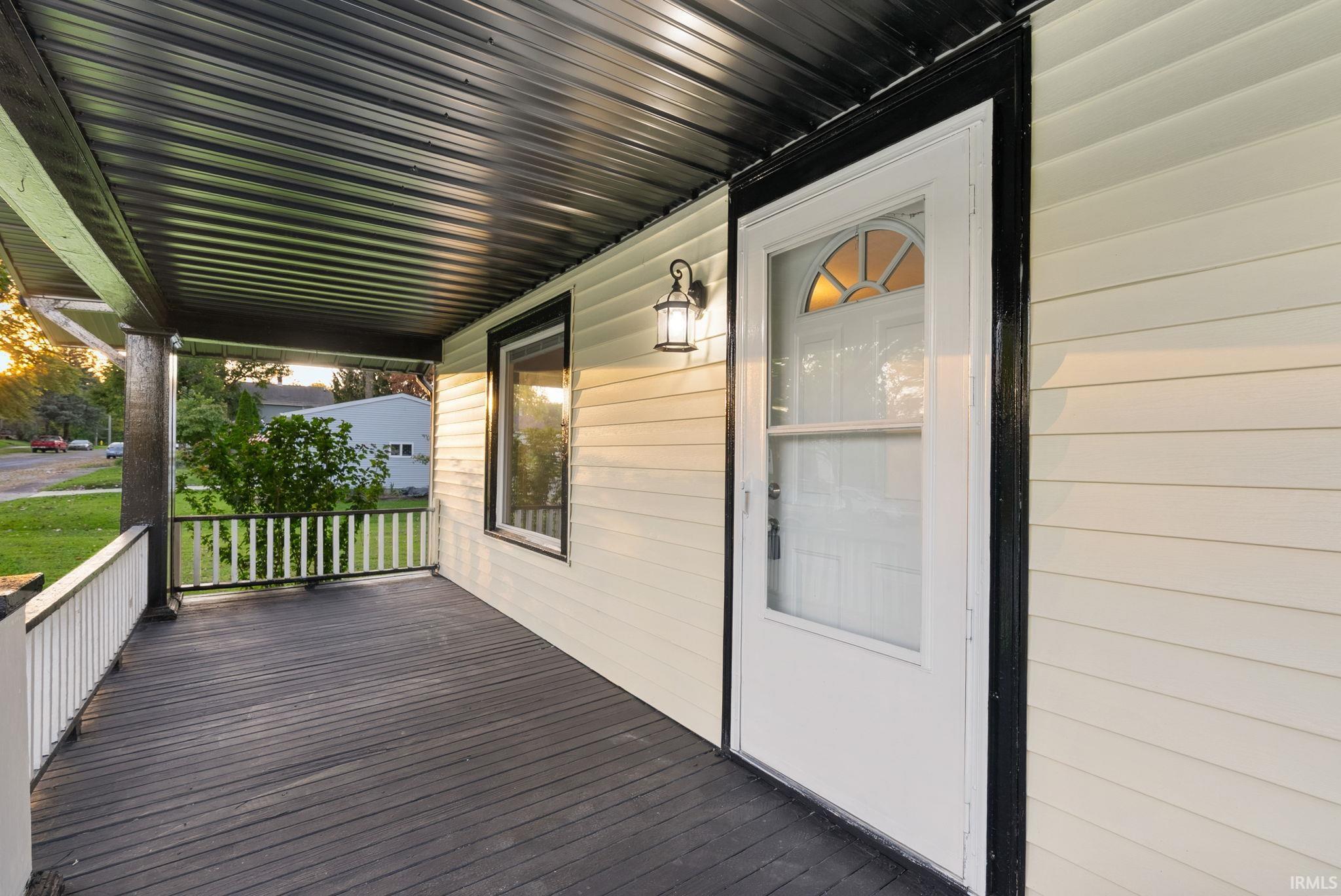 Covered front porch featuring dark-finished wood decking, contrast horizontal siding, black metal railing, and a wall-mounted exterior light fixture