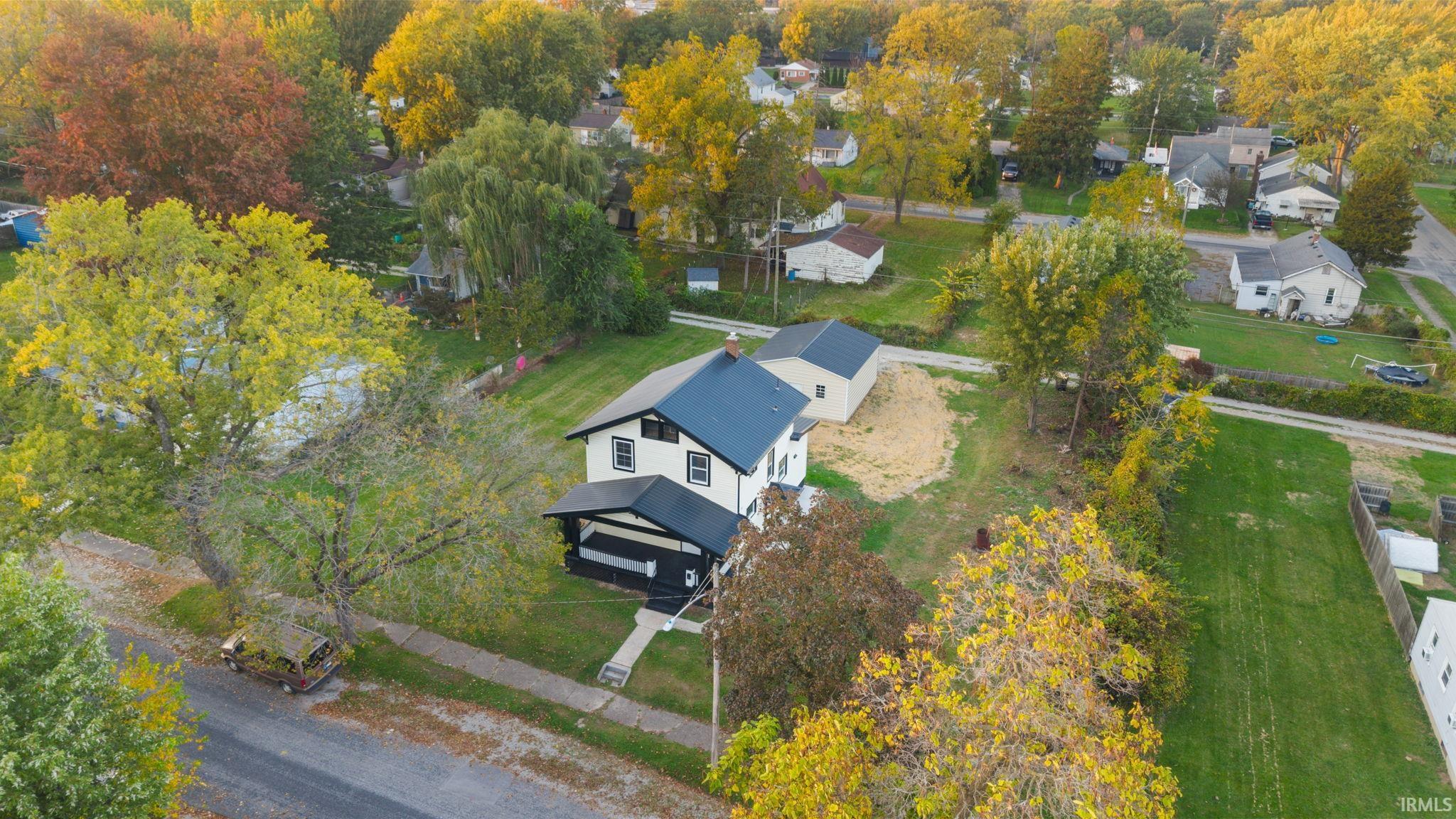 Aerial perspective of a multi-story residence featuring a dark-toned gable roof, covered front porch, exterior white siding, detached garage, and mature trees on the parcel