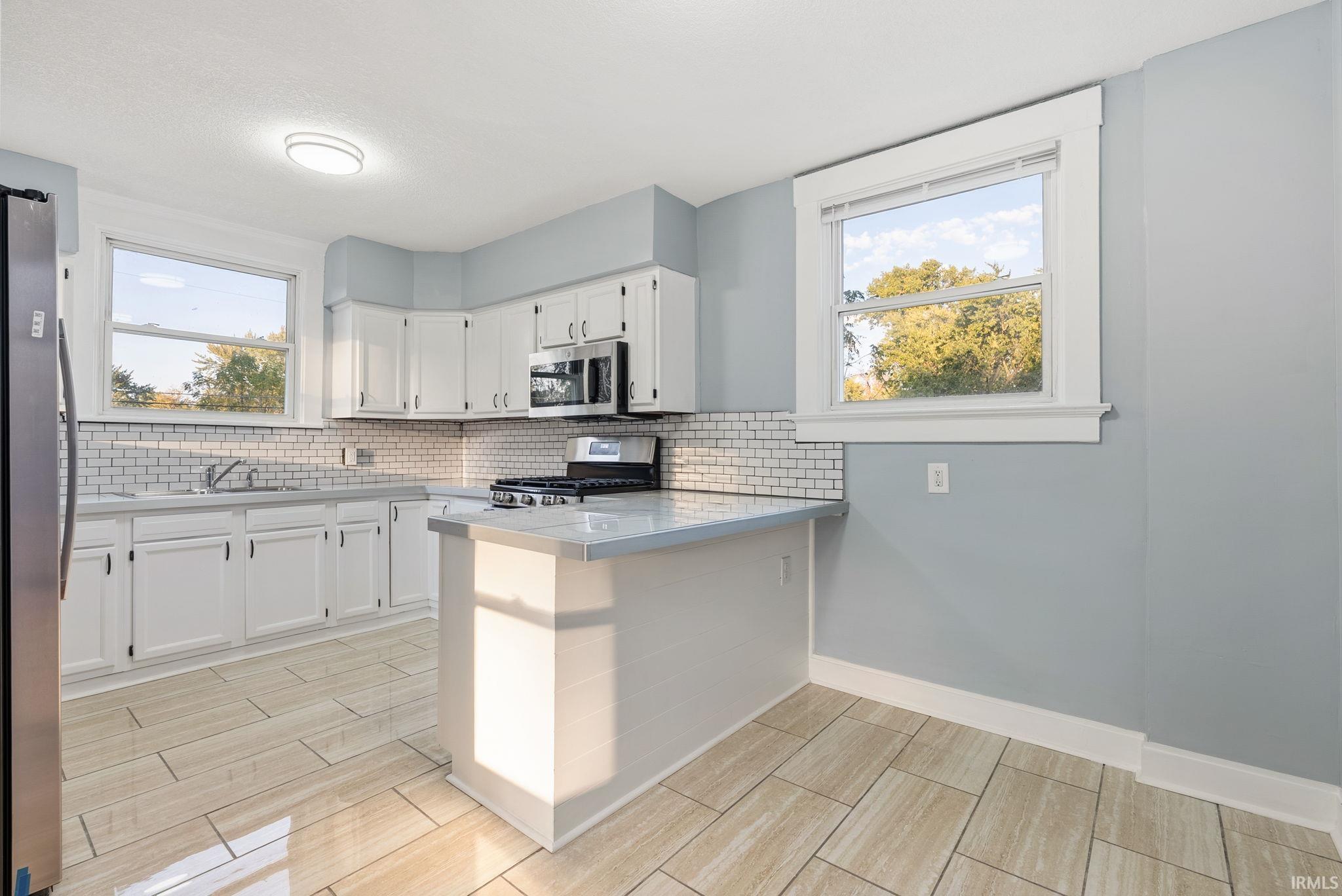 Kitchen featuring white cabinetry, light-toned tile flooring, subway tile backsplash, stainless steel appliances, and a peninsula with a solid-surface countertop