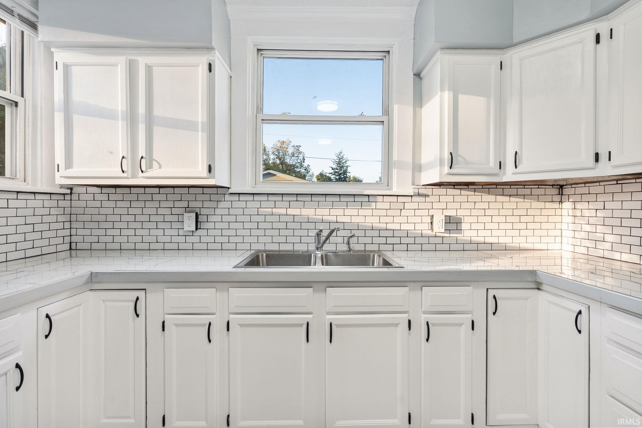 Kitchen featuring white shaker-style cabinetry, white subway tile backsplash, stainless steel dual-basin sink, chrome faucet, and light-toned solid surface countertops