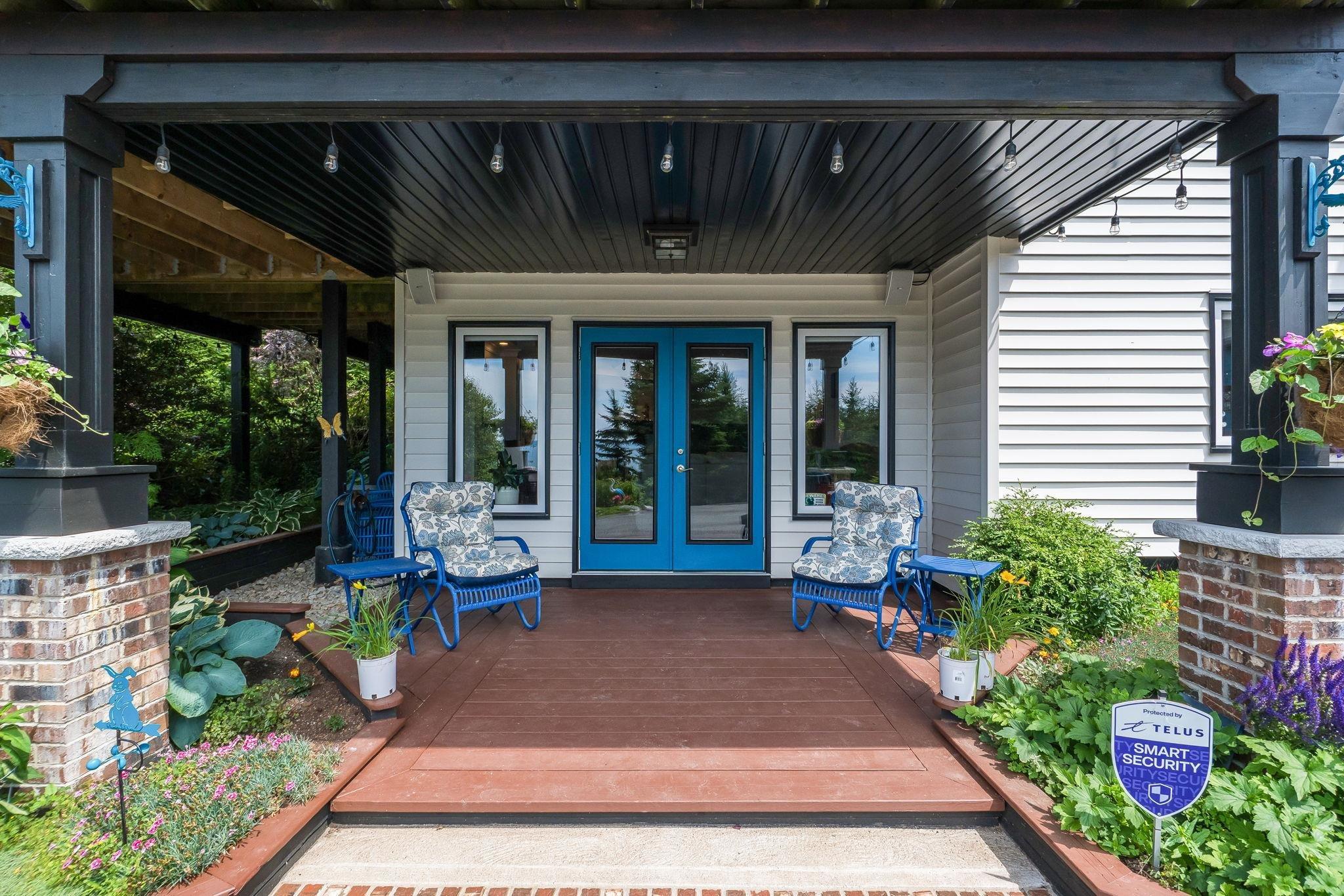 116 Whitecap Ridge, Black Point, NS (MLS® 202618413) — Covered front porch featuring a dark wood-finish ceiling, brick-clad columns, white horizontal siding, and a vibrant blue double door entry with glass panels
