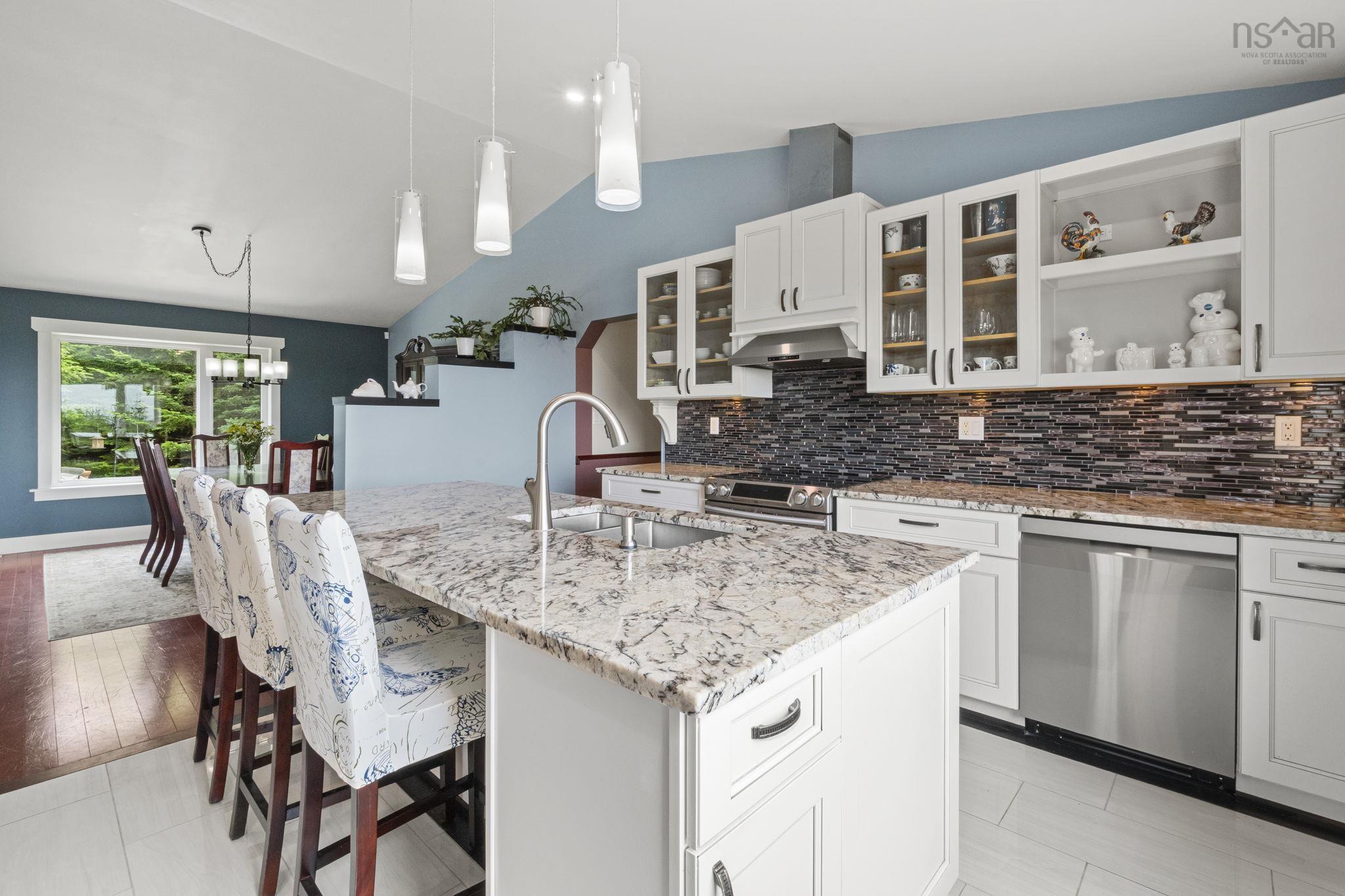 116 Whitecap Ridge, Black Point, NS (MLS® 202618413) — Kitchen featuring a central island with a granite countertop and integrated sink, white cabinetry with glass-front uppers, a mosaic tile backsplash, and stainless steel appliances