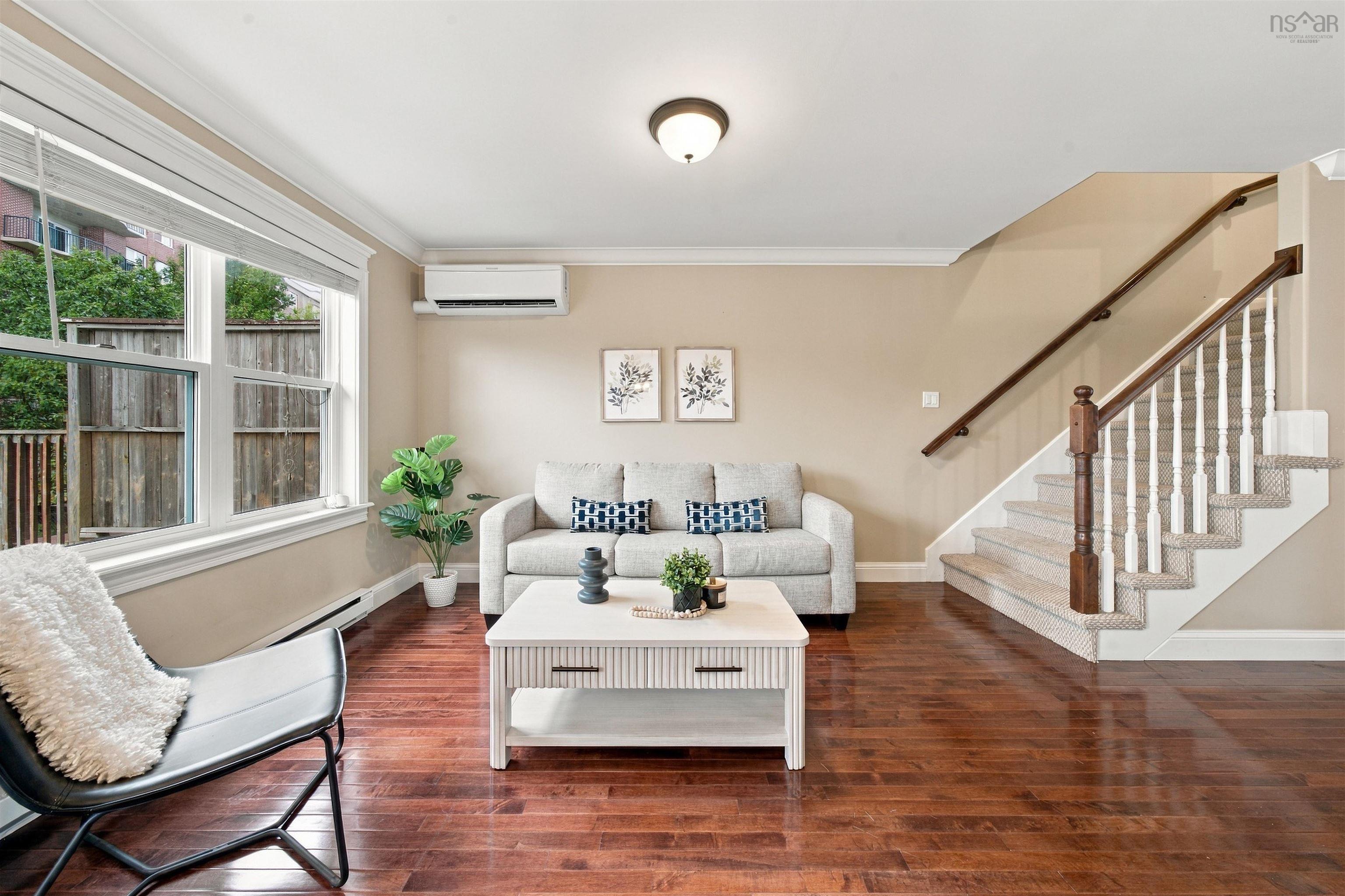 150 Transom Drive, Halifax, NS (MLS® 202625460) — Bright open-concept living area featuring hardwood flooring, large multi-pane windows with natural light, a white mantel fireplace, and a curved staircase with wood railings and white balusters