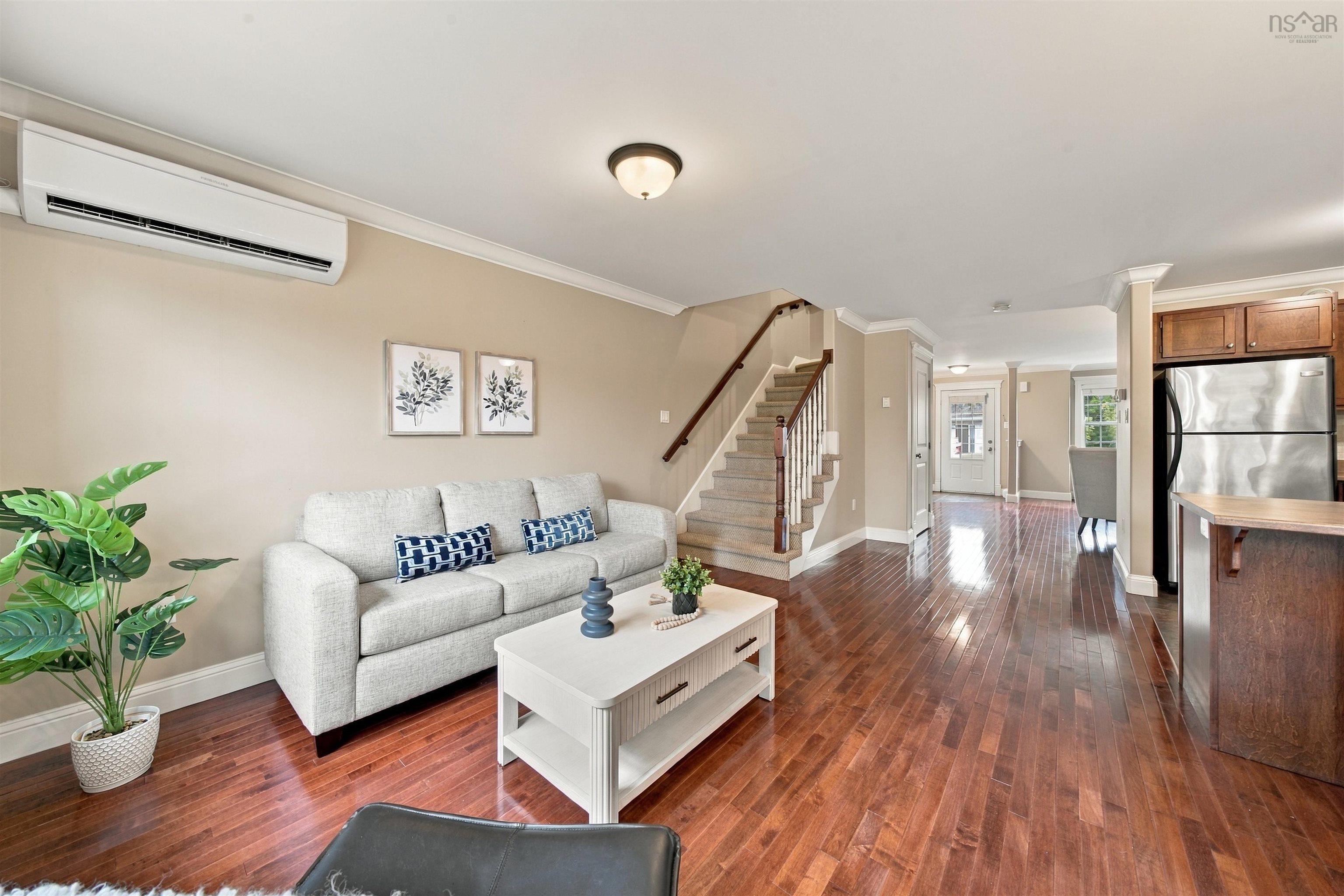 150 Transom Drive, Halifax, NS (MLS® 202625460) — Bright open-concept living area featuring hardwood flooring throughout, a white upholstered sofa with blue accent pillows, a white coffee table with storage, and a staircase with wooden treads and metal railings