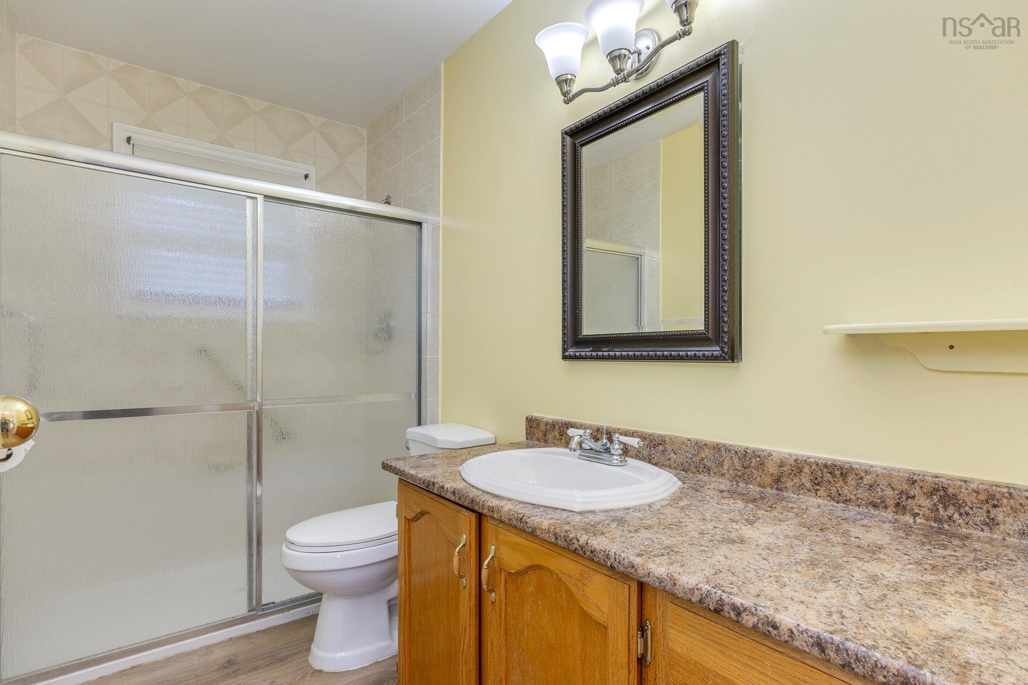 128 Doherty Drive, Lawrencetown, NS (MLS® 202625891) — Bathroom featuring a wood-finish vanity with a stone-composite countertop, integrated sink, shower with sliding glass doors, and three-light vanity fixture