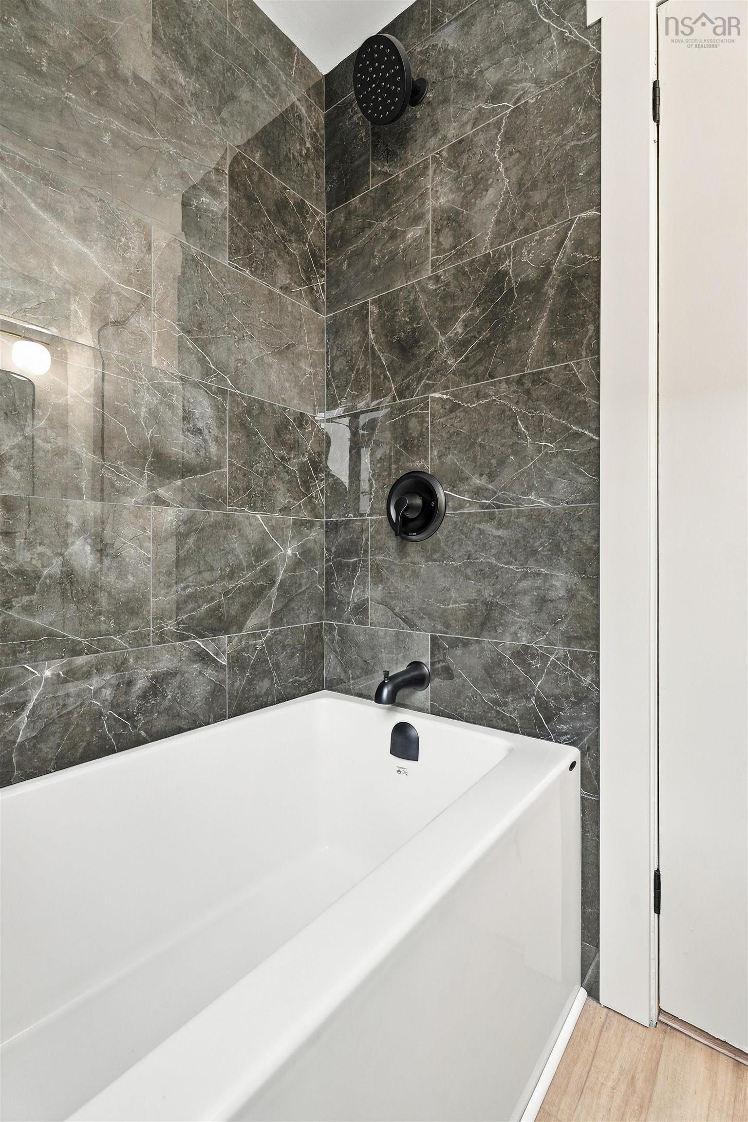 74 1/2 Belle Vista Drive, Dartmouth, NS (MLS® 202625912) — Deep-soaking bathtub with a white finish, surrounded by large-format gray tile wall accents, featuring matte black shower head, faucet, and hardware