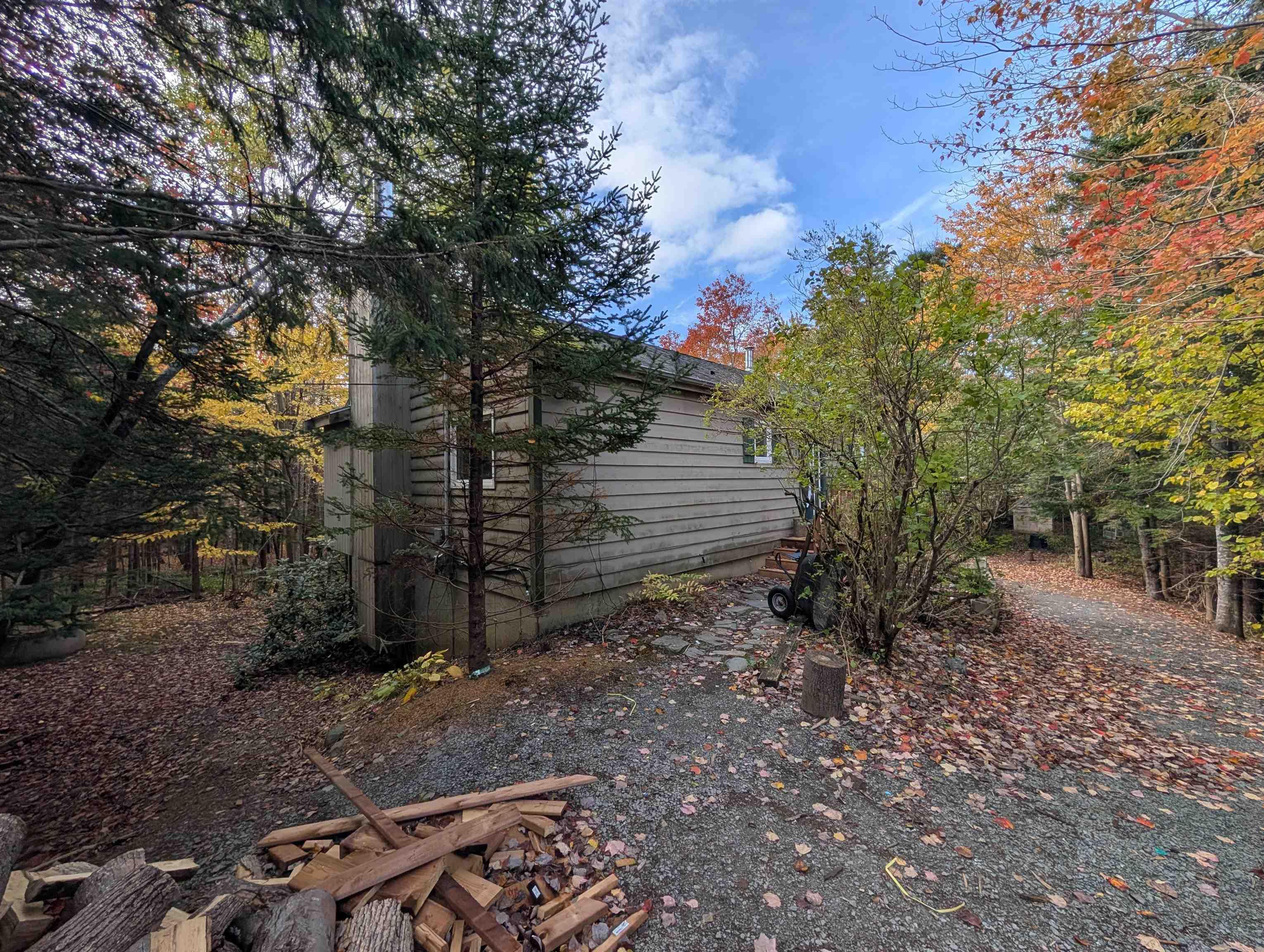19 Foster Avenue, Fall River, NS (MLS® 202625937) — Exterior featuring horizontal wood siding, a stone chimney, and a gravel driveway surrounded by mature trees