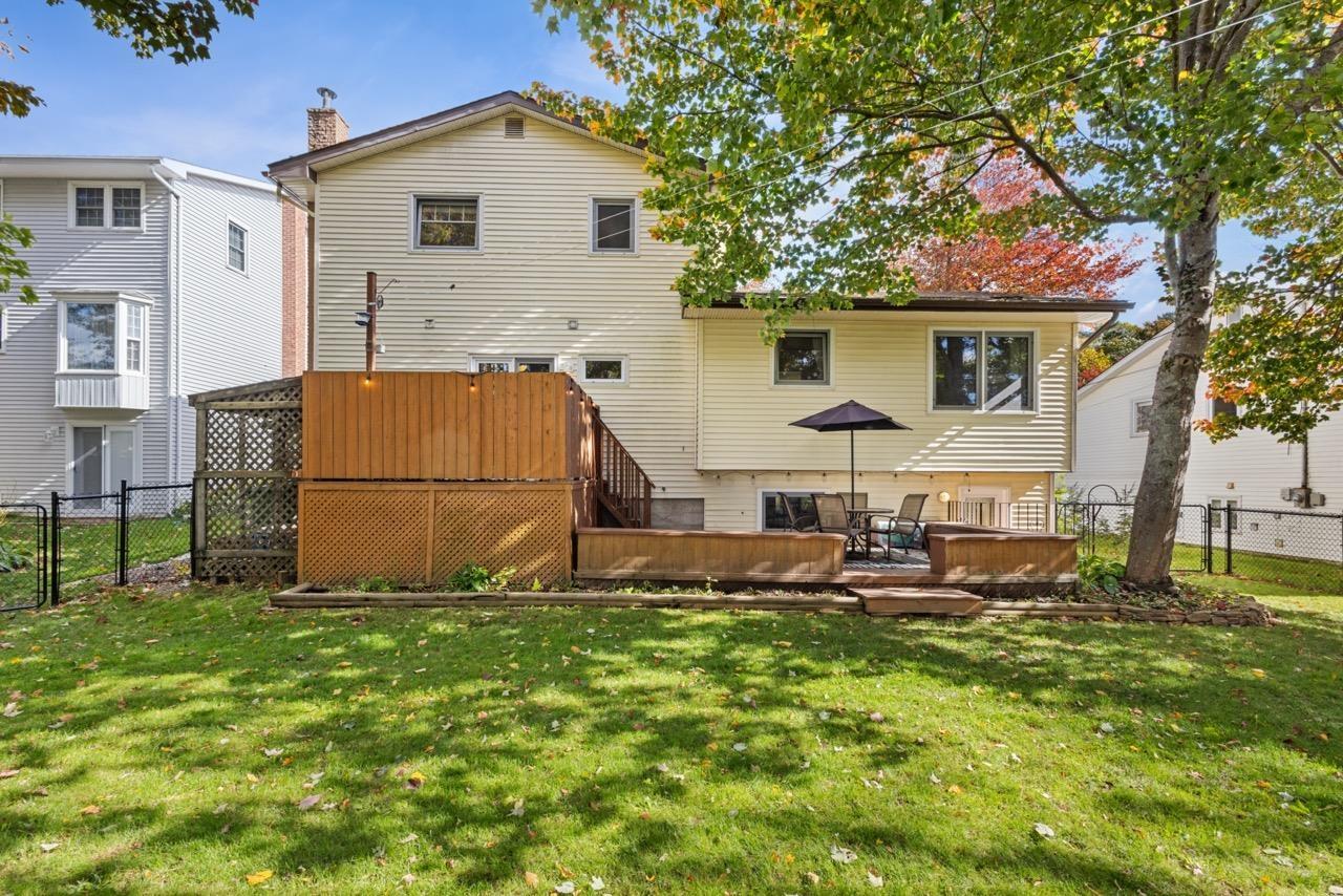 24 Rossi Drive, Dartmouth, NS (MLS® 202626410) — Rear exterior featuring light-colored siding, a multi-level wood deck, lattice privacy screening, and a fenced-in grass lawn with mature trees