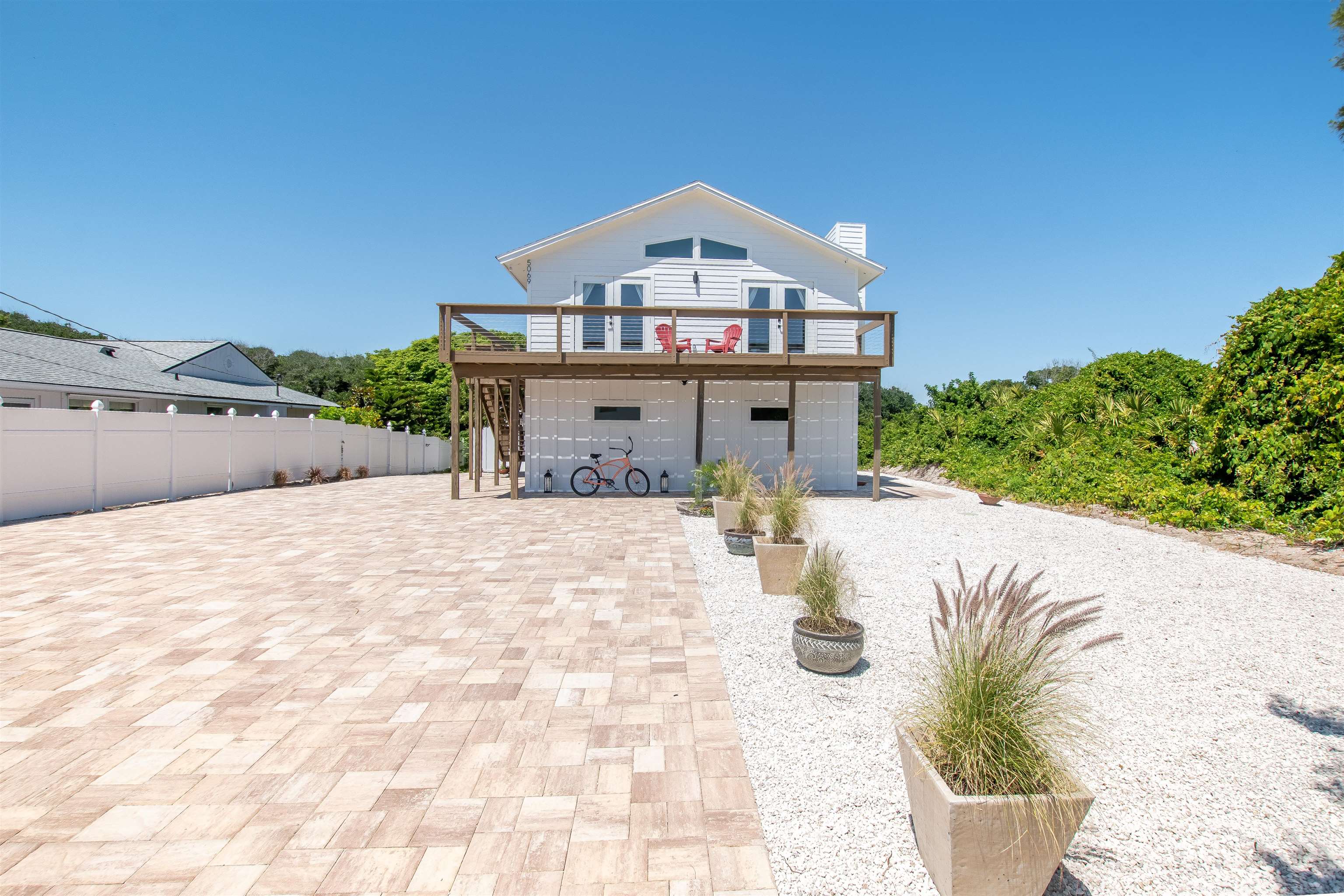 White exterior beach cottage with a second-story wood deck, a light-colored paver driveway, and a white fence Front of house!