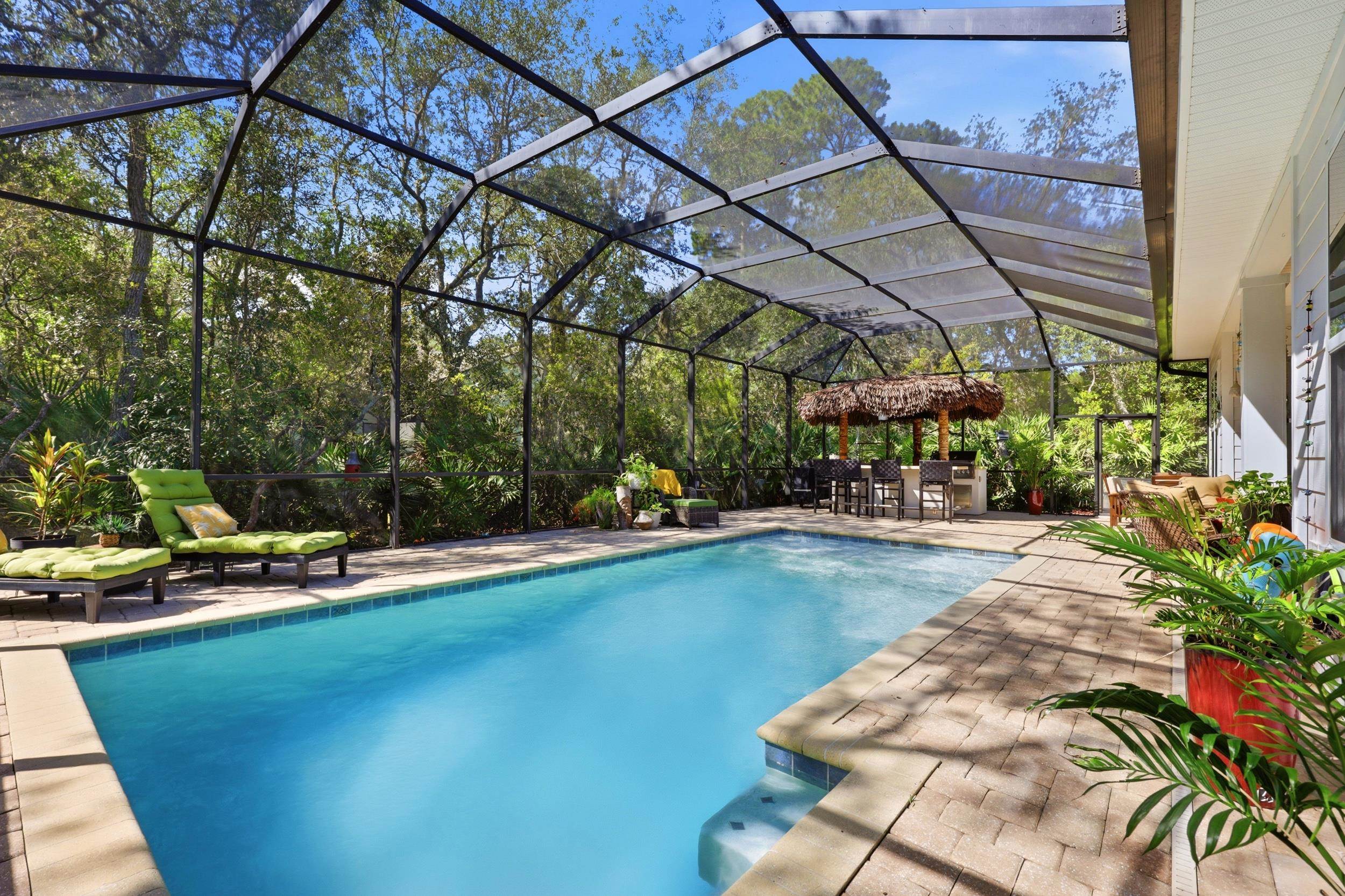 Screened-in pool area featuring a built-in outdoor bar with a thatched roof, paver deck, and mature perimeter landscaping Pool