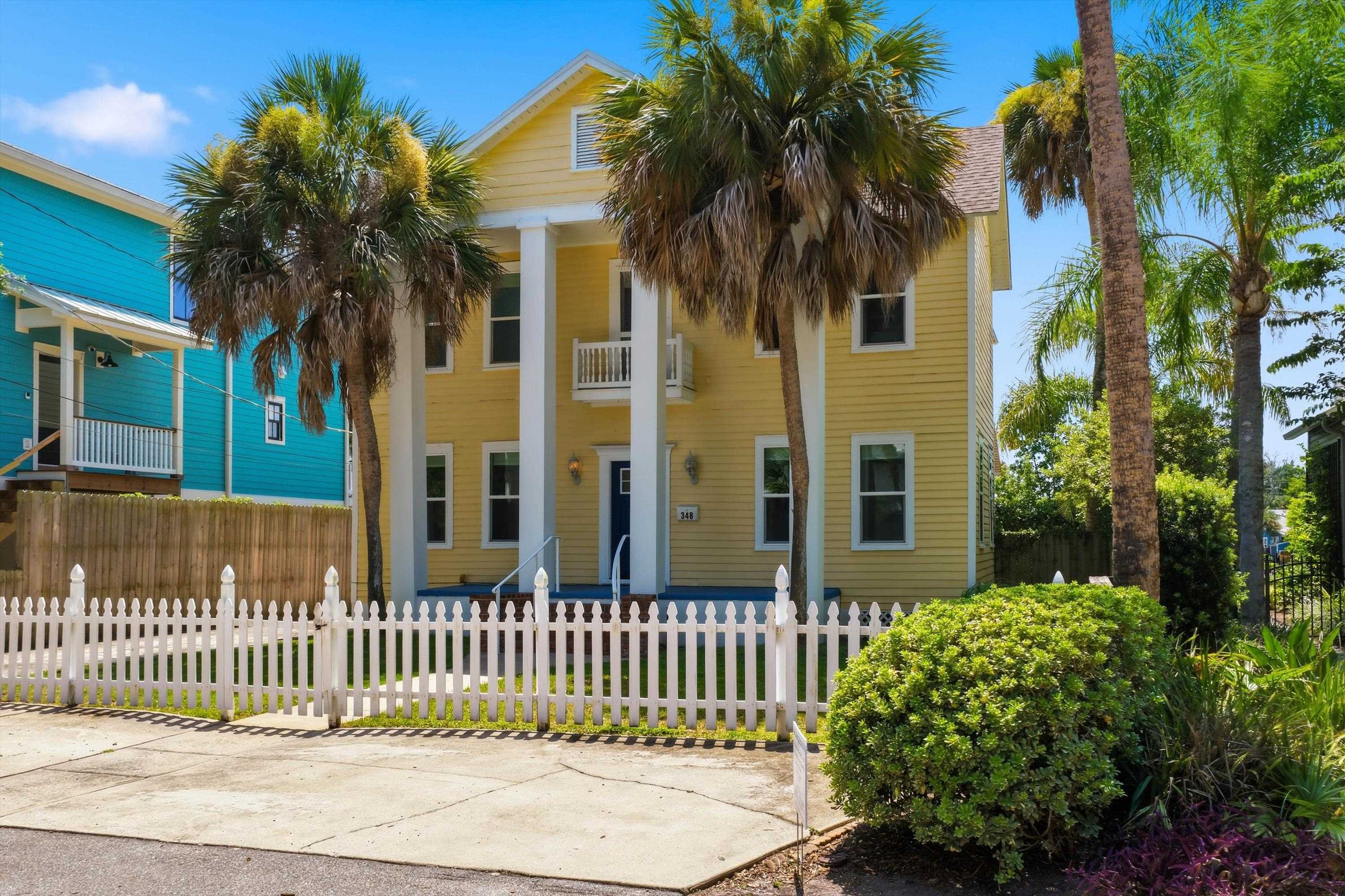 Two-story yellow residence featuring a prominent columned entrance with an upper-level balcony, fronted by a white picket fence Front Of Structure