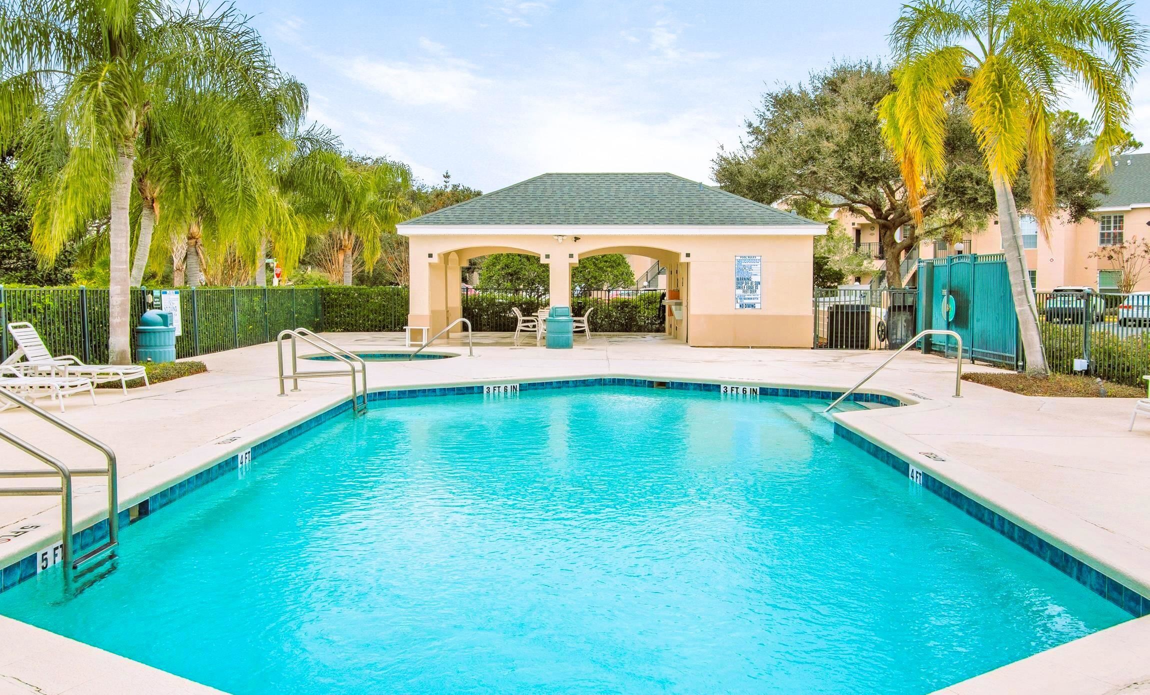 Community swimming pool with an adjacent spa, surrounded by a paved deck, a shaded cabana, and mature palm trees Pool