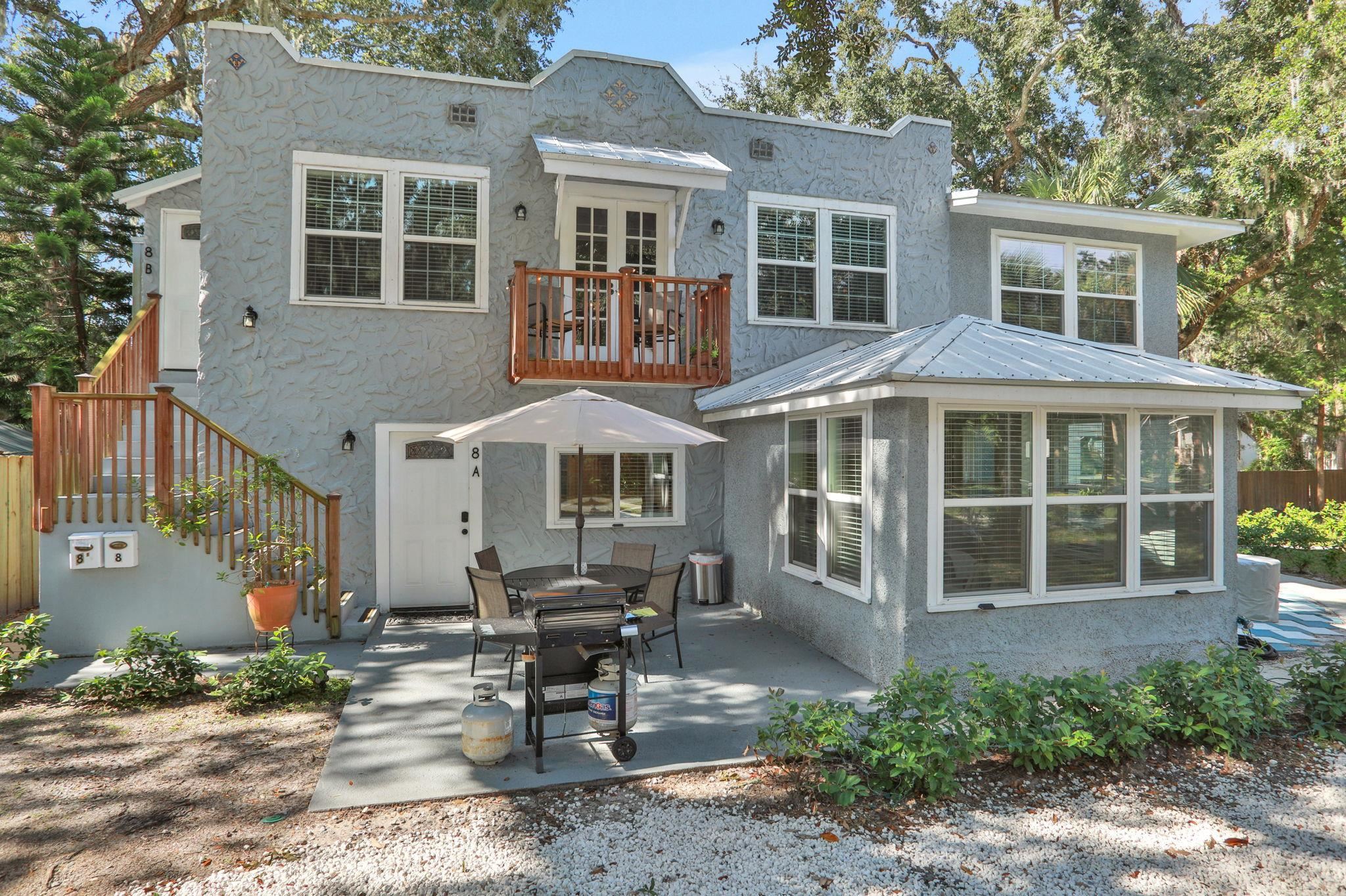 Stucco exterior with multiple white-framed windows, a private second-story balcony with wood railings, an exterior staircase with wood railings, a sunroom with a metal roof, and a ground-level patio area Back Of Structure