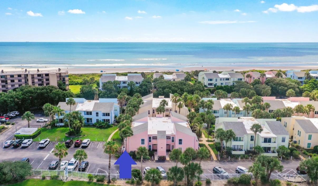 Coastal community setting with a multi-story building, adjacent parking, and mature palm trees, positioned near the oceanfront Aerial View