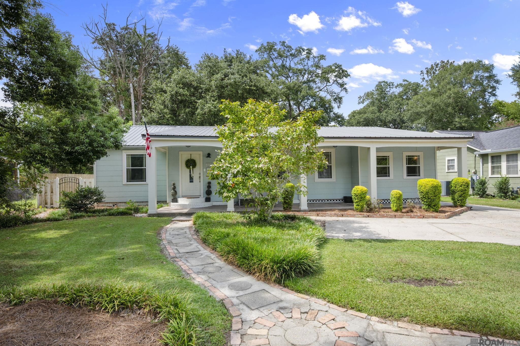 Pale blue single-story home with a covered porch, mature trees, and a curved stone walkway.